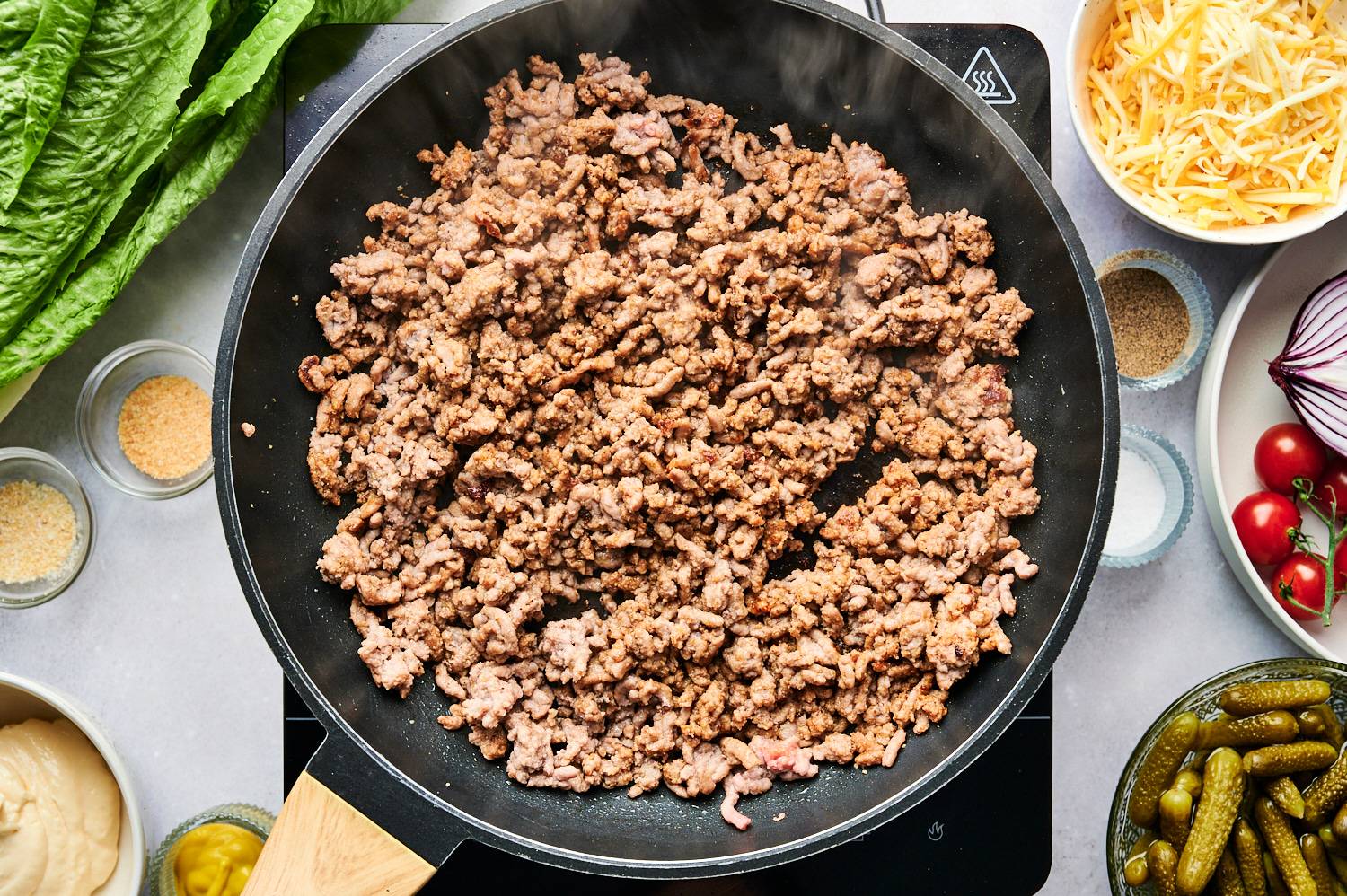 Burger seasoned ground beef in a skillet with burger toppings on the size.