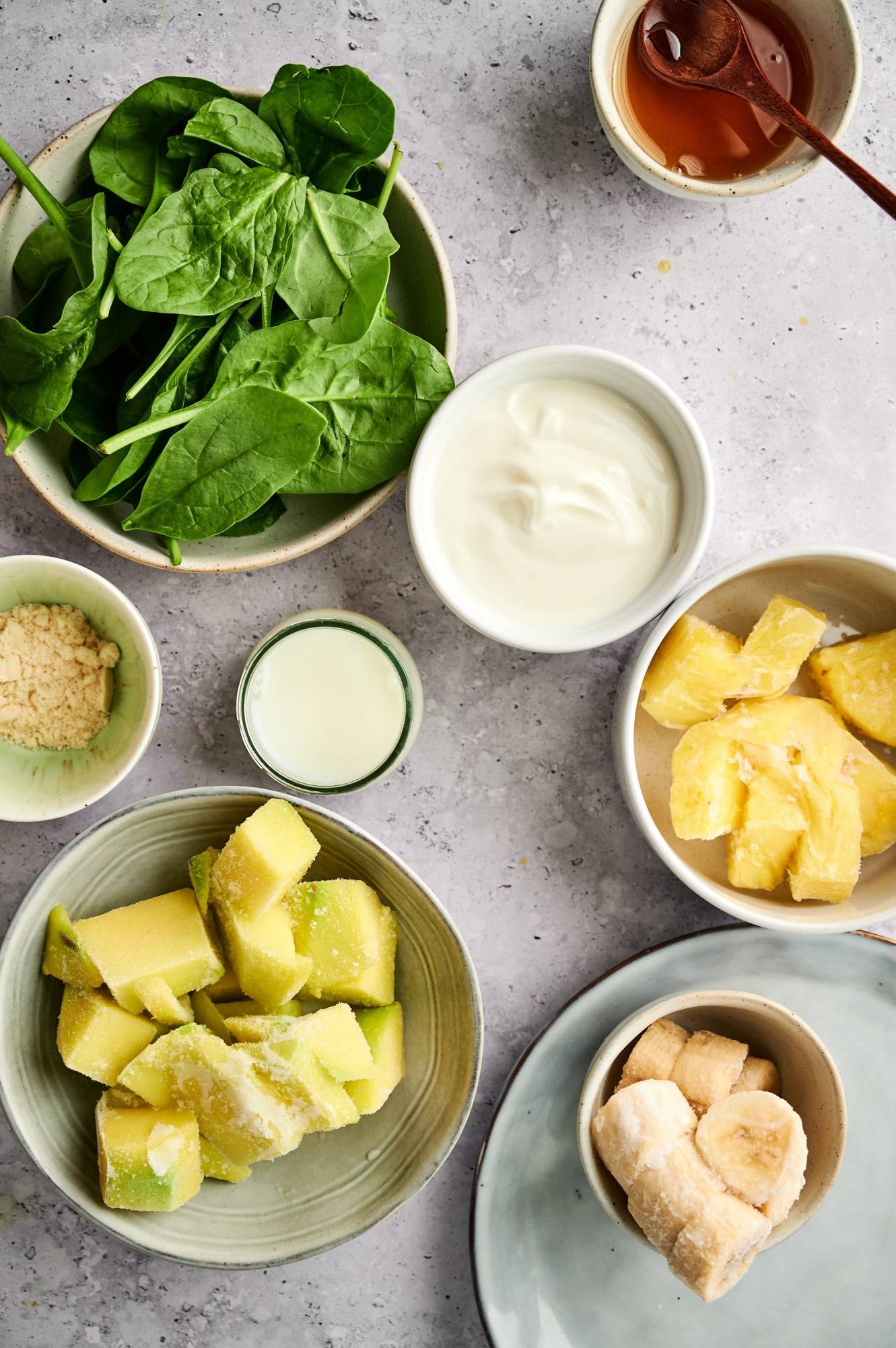 Ingredients for green smoothie bowls including frozen mango, pineapple, banana, baby spinach, milk, yogurt. and protein powder.