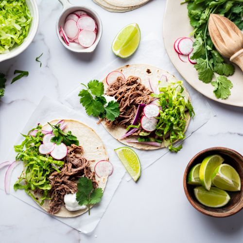 Crockpot tri-tip shredded tacos with red onion, cilantro, lettuce, and radishes served on corn tortillas.