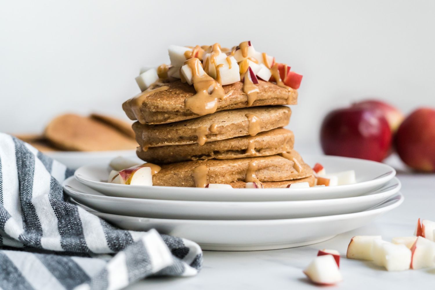 Apple cinnamon pancakes stacked on a plate with melted peanut butter and chopped apples.