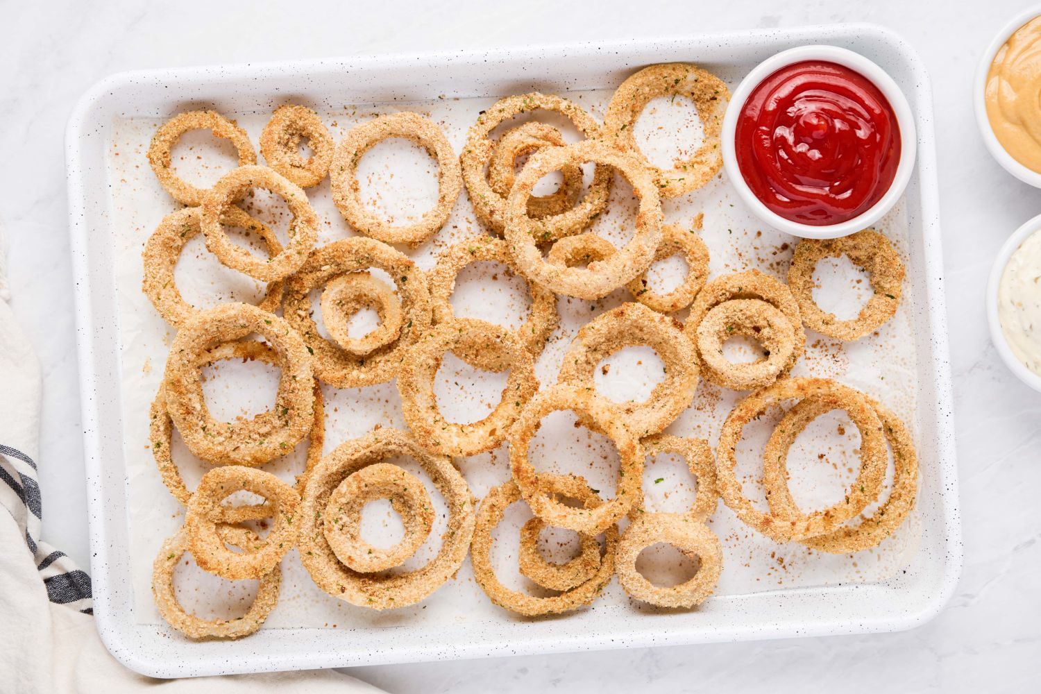 Baked onion rings with a golden brown breadcrumb coating on a baking sheet with ketchup on the side.