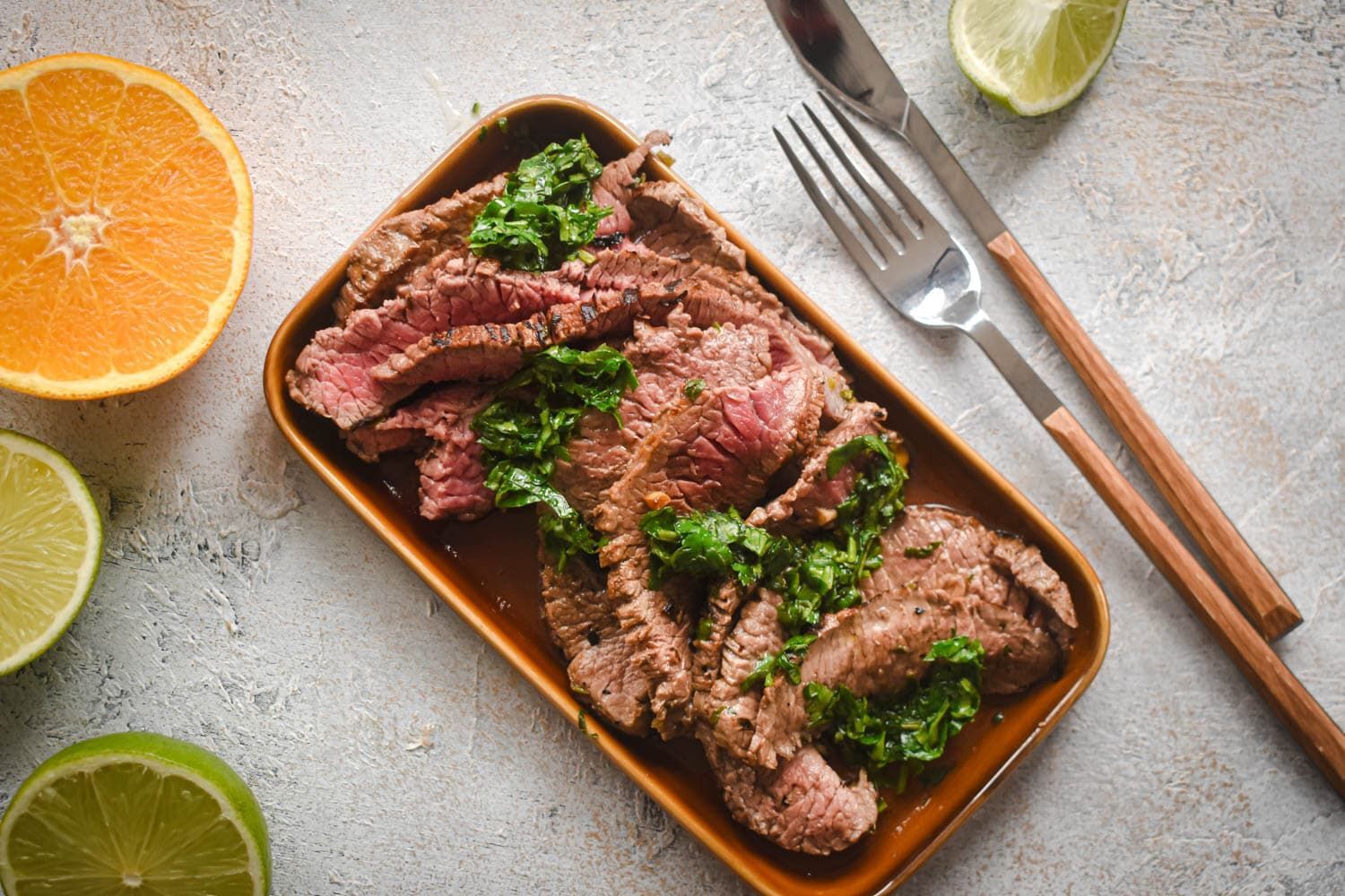 Carne asada marinated in citrus juices sliced thin on a plate with cilantro, limes, and orange slices.
