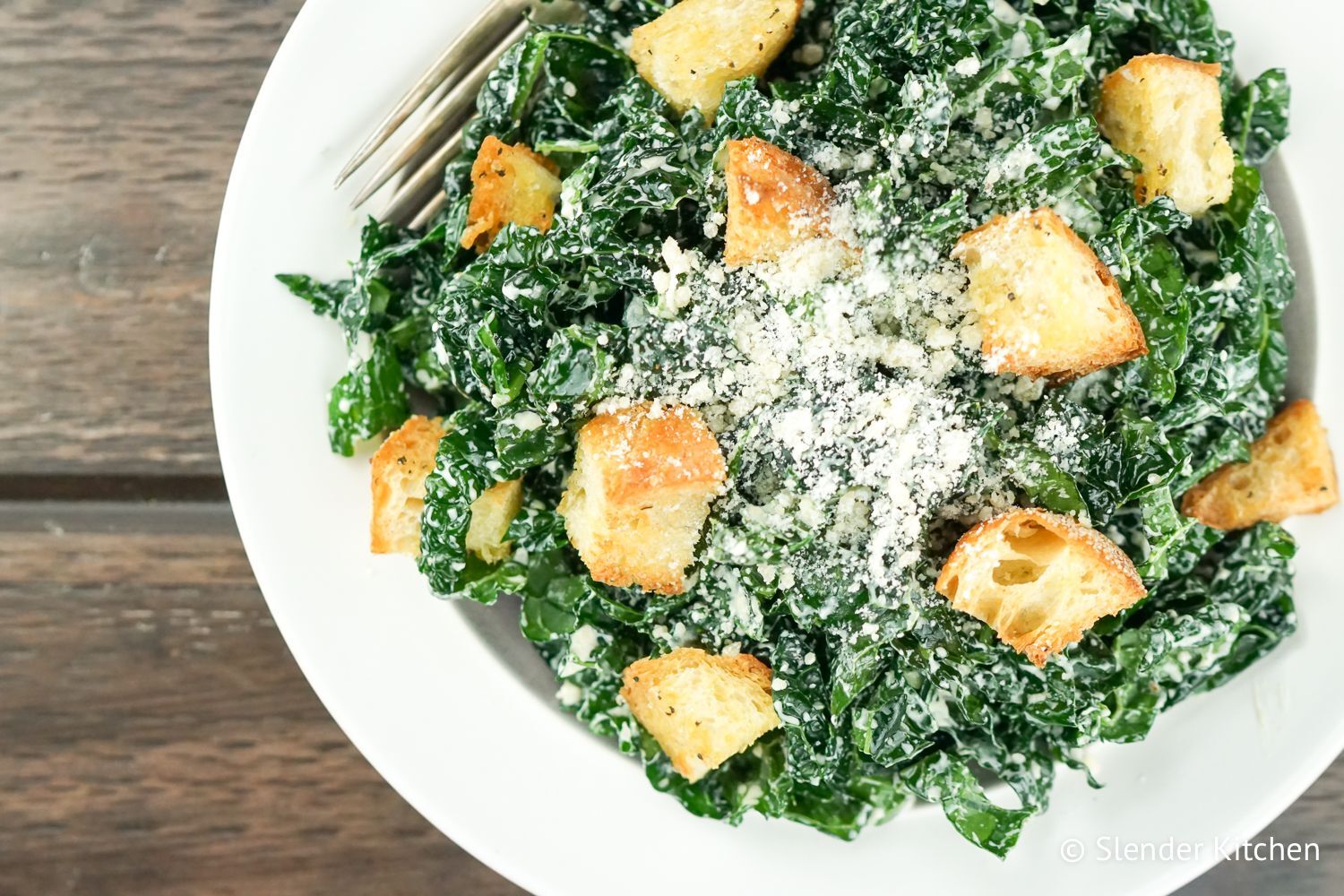Kale caesar salad with croutons and Parmesan cheese on a plate with a fork.