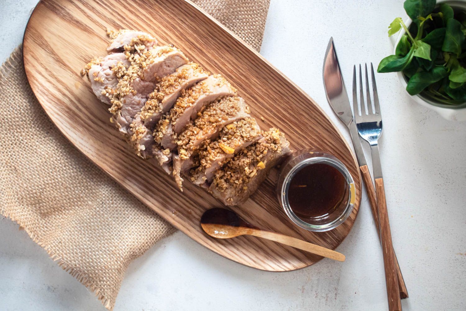 Honey pork tenderloin coated with sesame seeds sliced on a wooden board with a soy garlic marinade on the side.