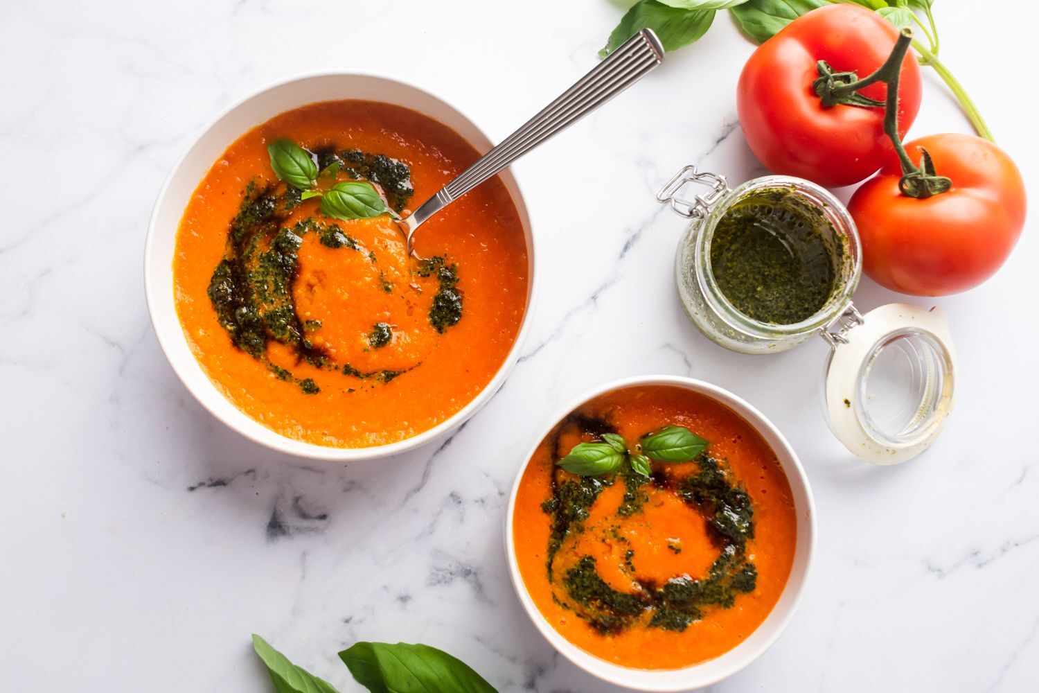 Roasted tomato basil soup in two bowls with pesto and fresh basil leaves.