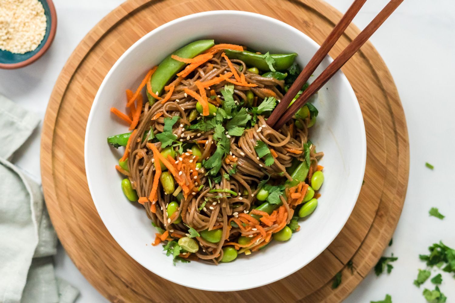 Asian noodle bowls with soba noodles, sesame dressing, carrots, edamame, and snap peas in a white bowl with chopsticks.