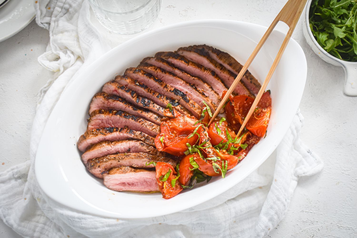 Balsamic flank steak sliced and served with roasted tomatoes in a white plate with wooden tongs.