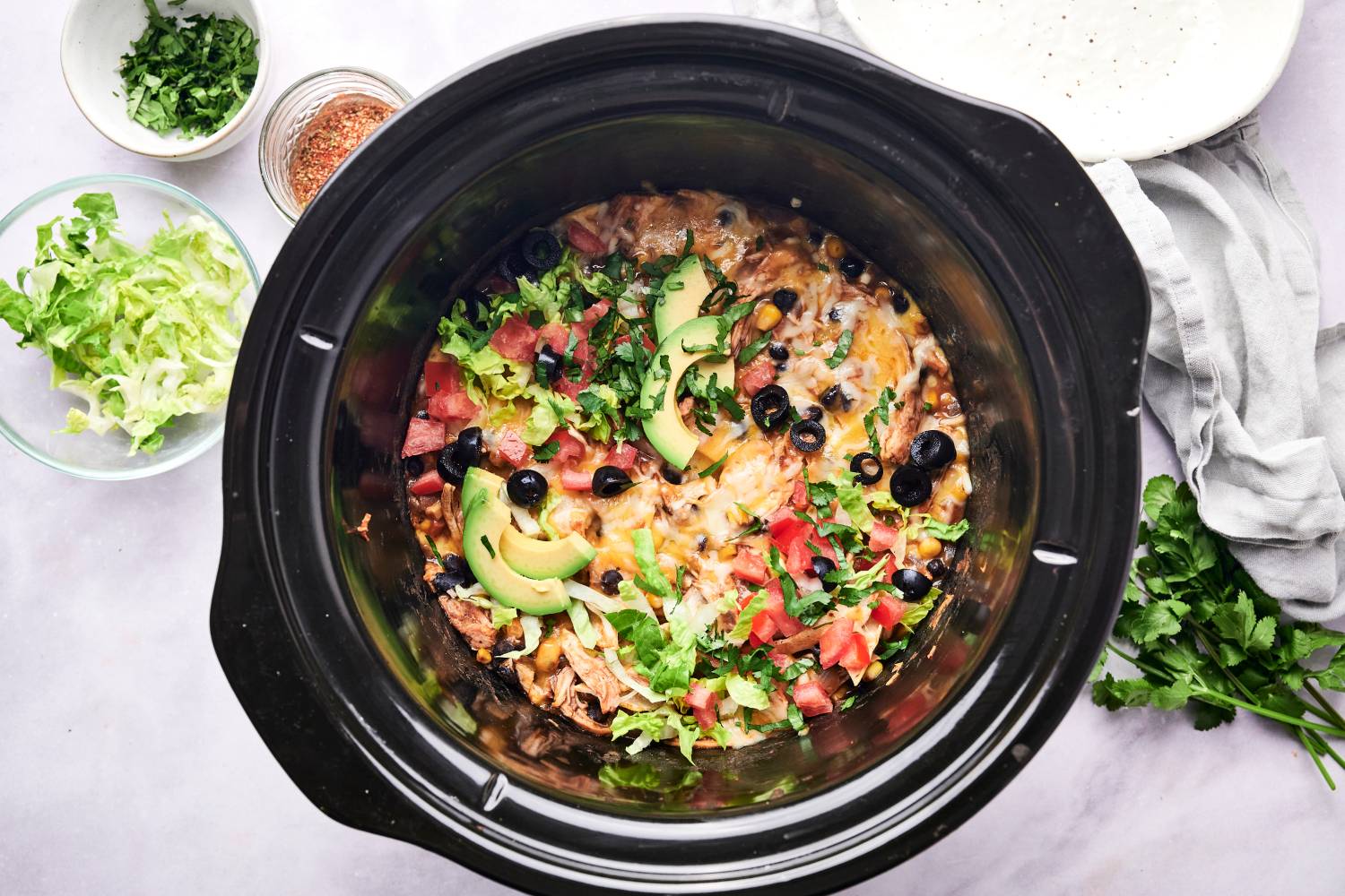 Slow cooker chicken enchilada casserole in a crockpot with avocado, black olives, lettuce, and tomatoes.