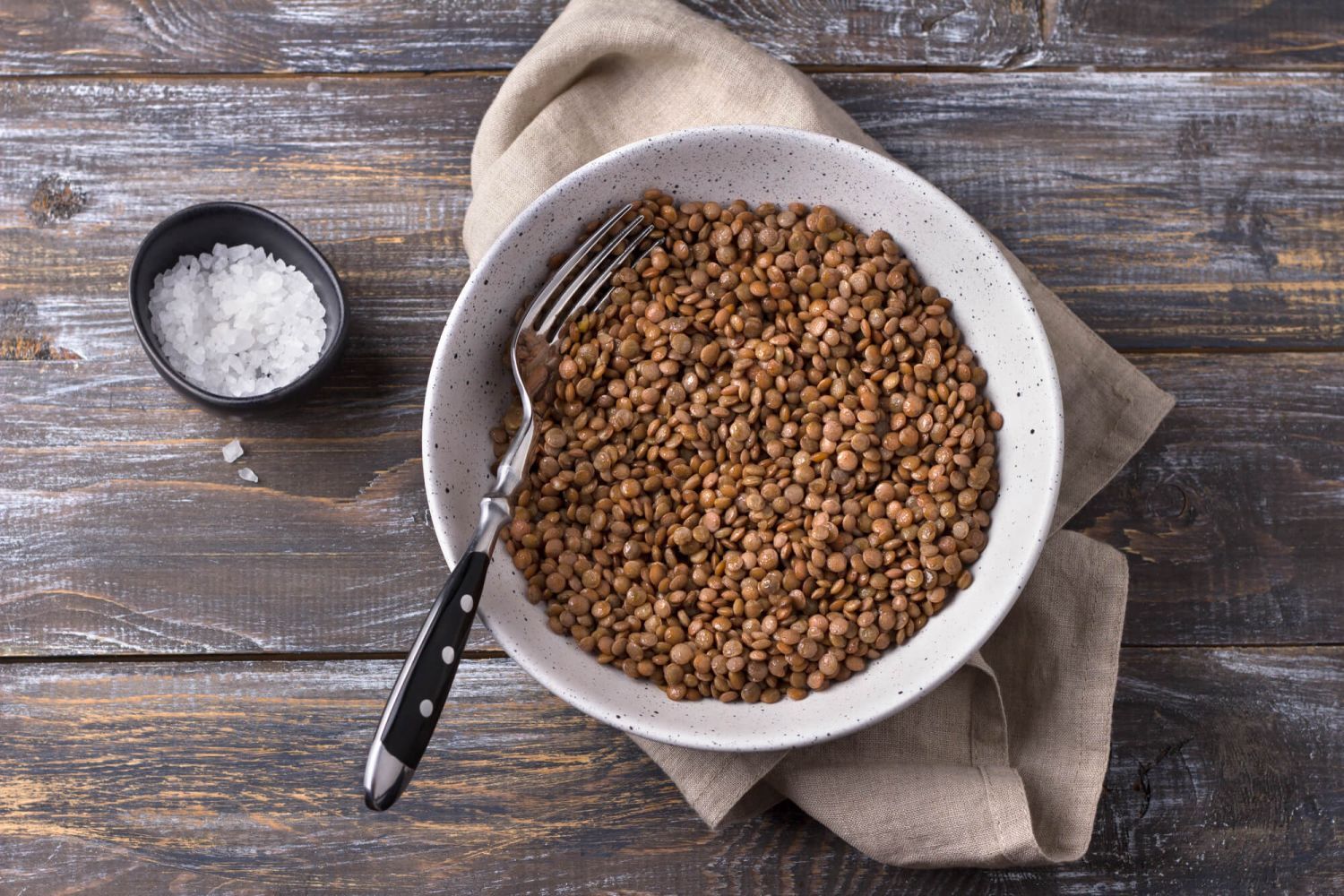 Cooked brown lentils in a white bowl with a napkin and fork.