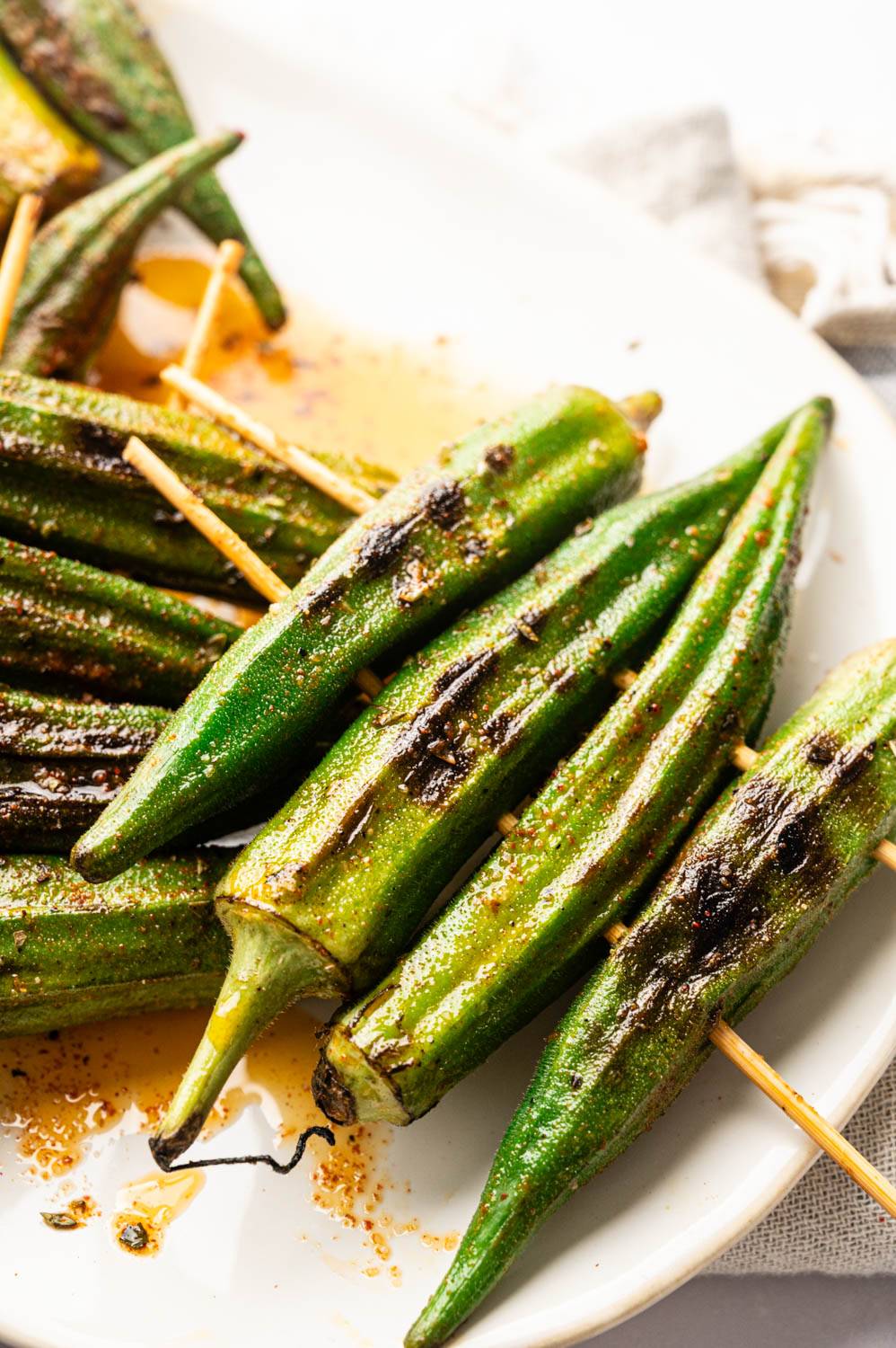 Close-up of grilled okra skewers with charred grill marks and Cajun seasoning, served on a white platter.