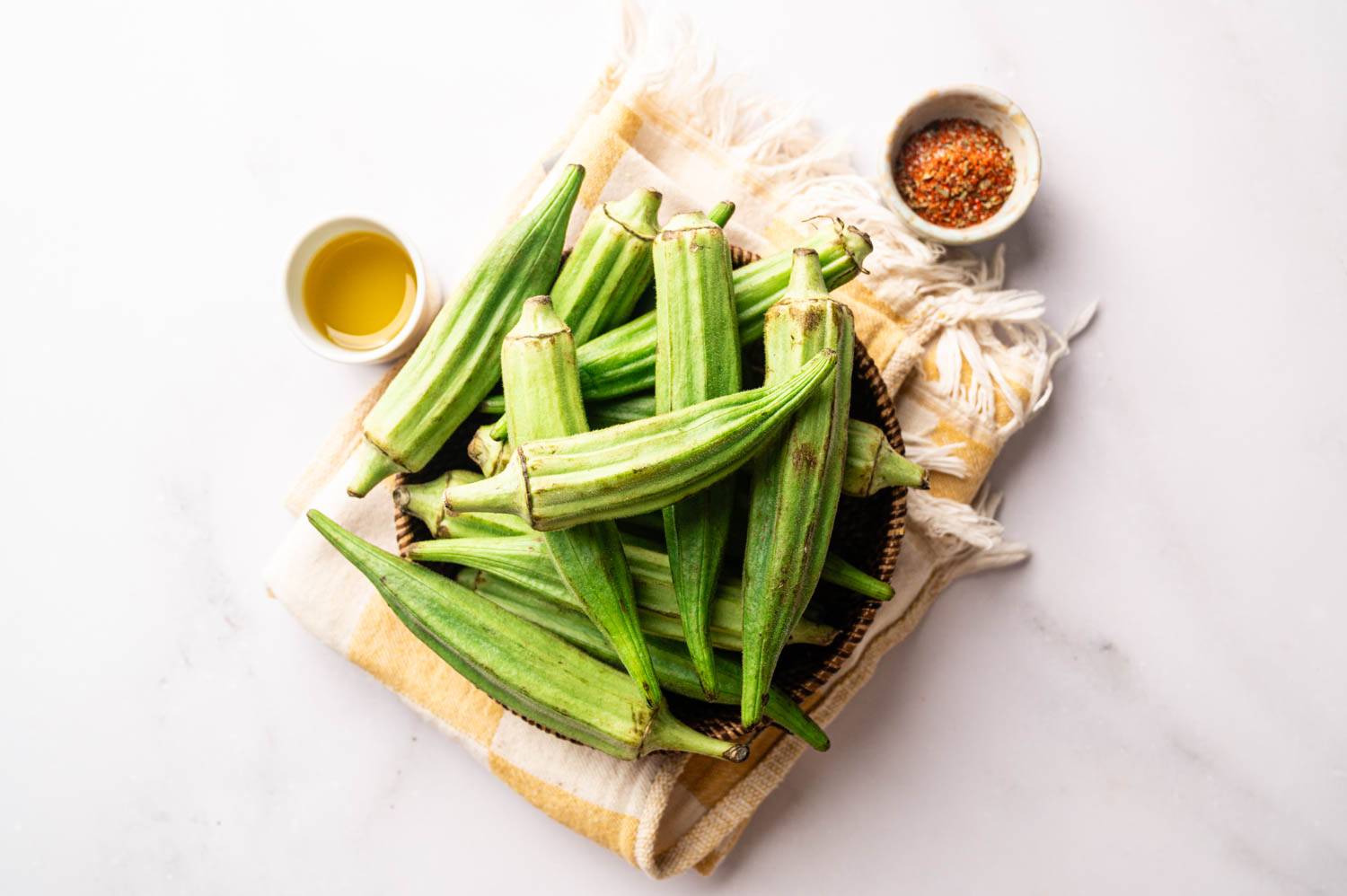 Fresh whole okra sits in a woven basket beside olive oil and Cajun seasoning, ready to be prepped for grilled okra skewers.