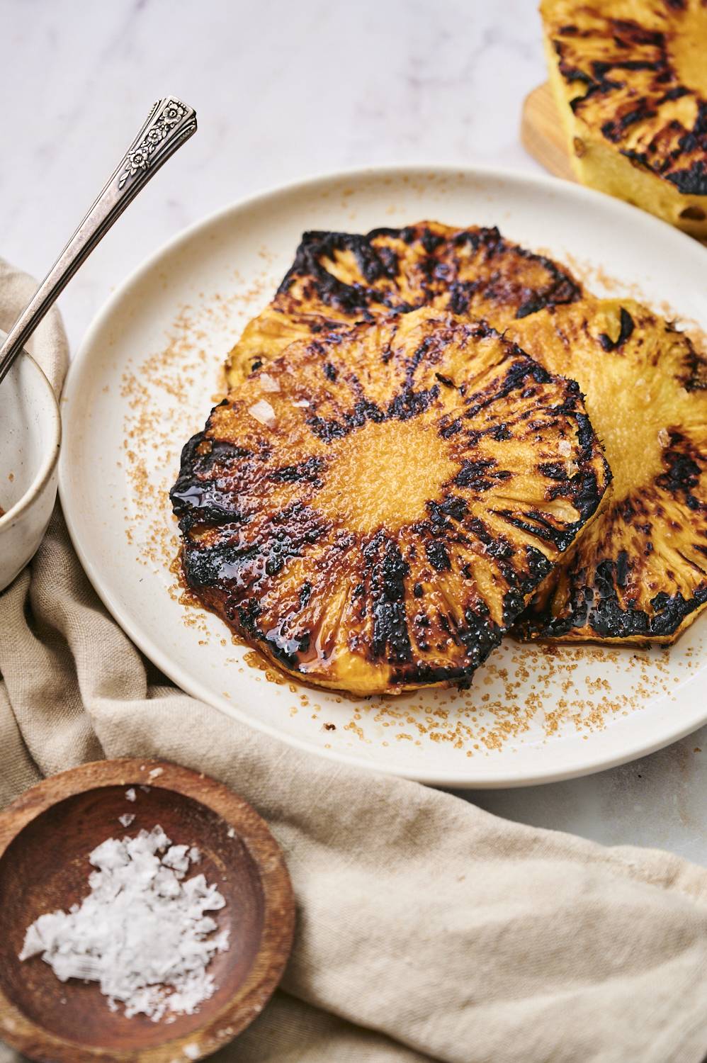 Grill pineapple slices on a white plate, top with sugar, a bowl of flakes of sea salt and silver spoon.