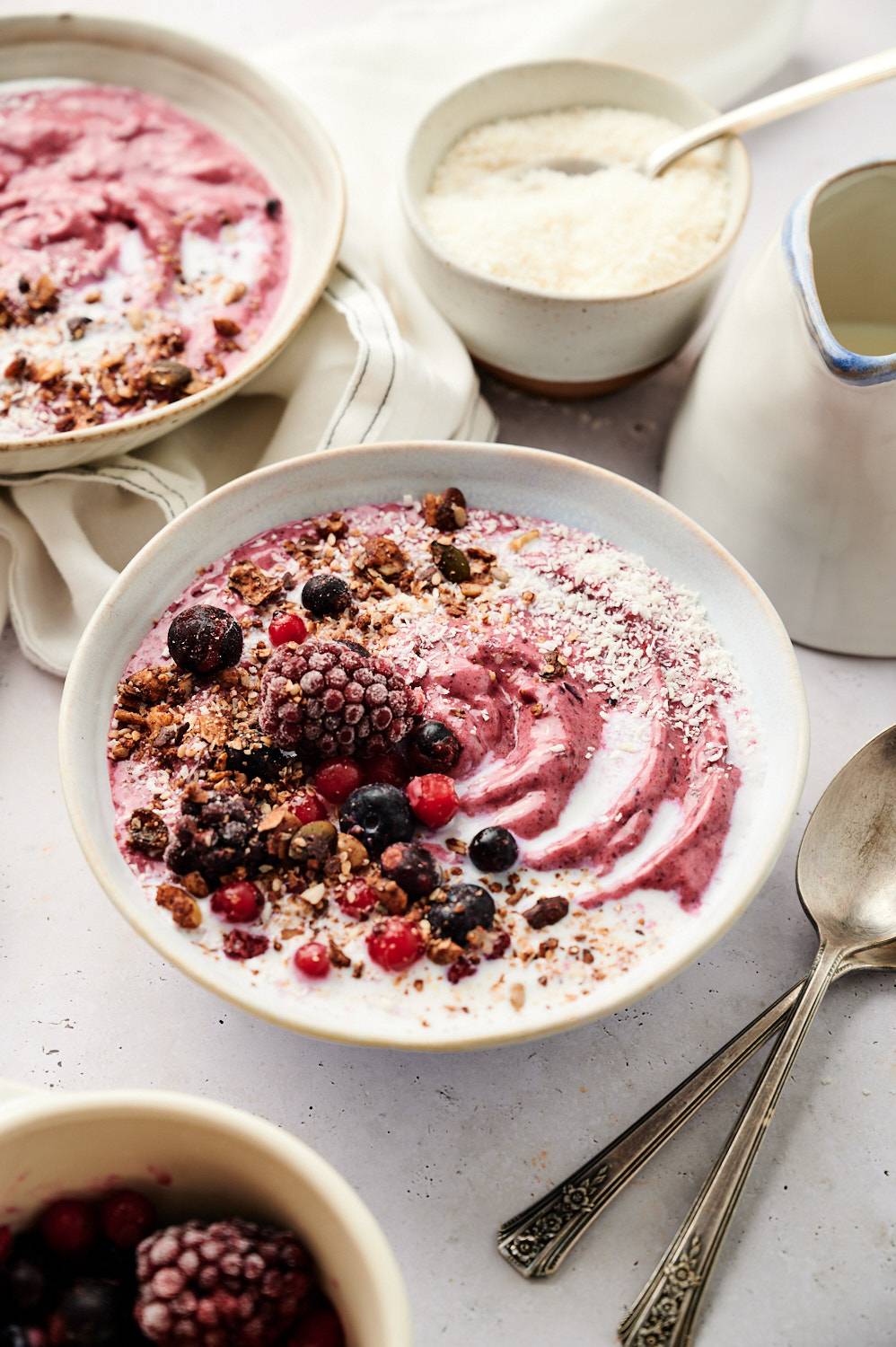 Acai bowls are topped with frozen berries, granola and coconut flakes, and are decorated with swirls of yogurt and milk in a ceramic bowl.