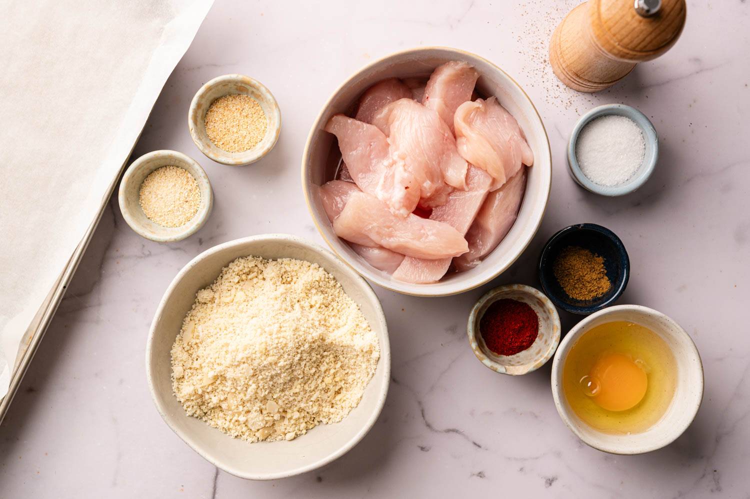 Raw chicken pieces, almond flour, egg, and spices are arranged on a marble countertop. A pepper grinder and parchment paper are visible.