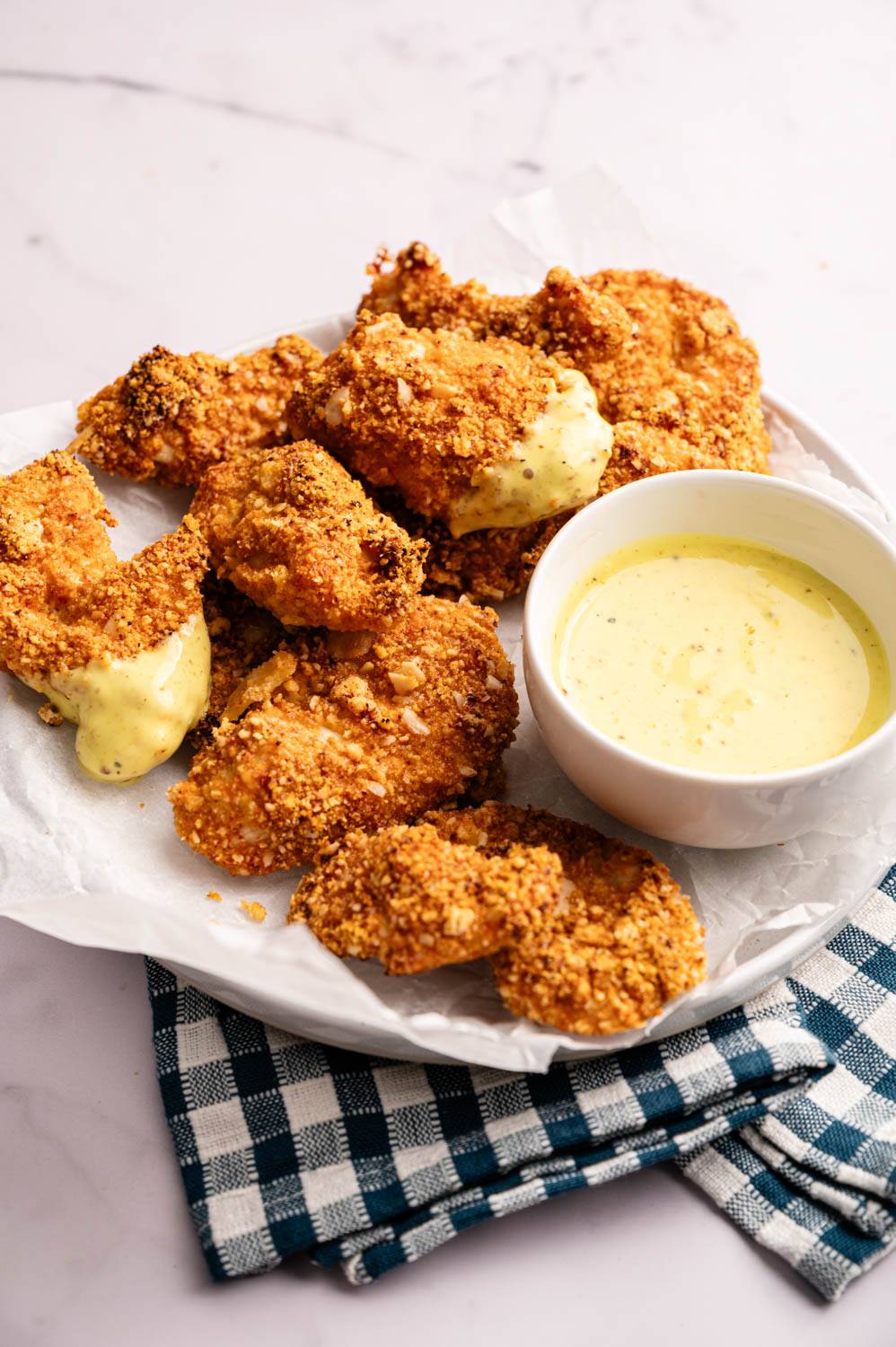 A plate of crispy, golden-fried shrimp coated in breadcrumbs sits on checkered cloth. Beside it, a cup of creamy yellow sauce, ready for dipping.