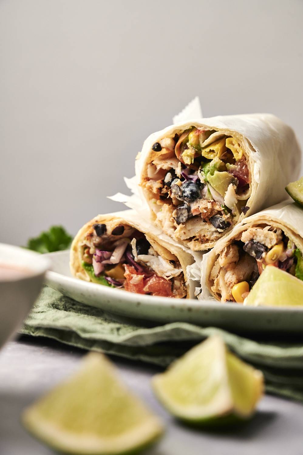 Stacked fish burritos filled with avocado, black beans, corn, and coleslaw on a plate with lime wedges.