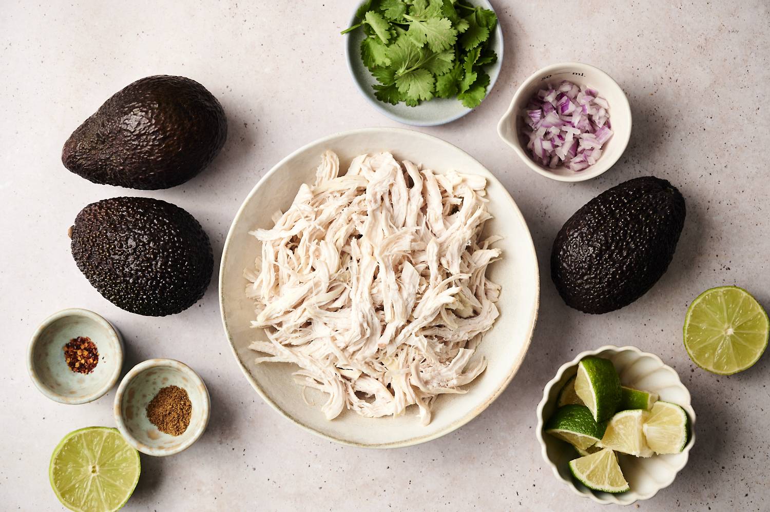 Flat ingredients of avocado chicken salad, including a bowl of chopped chicken, surrounded by whole avocado, chopped red onions, fresh coriander, lime wedges, half lime, ground spices and red pepper flakes and red pepper flakes. 