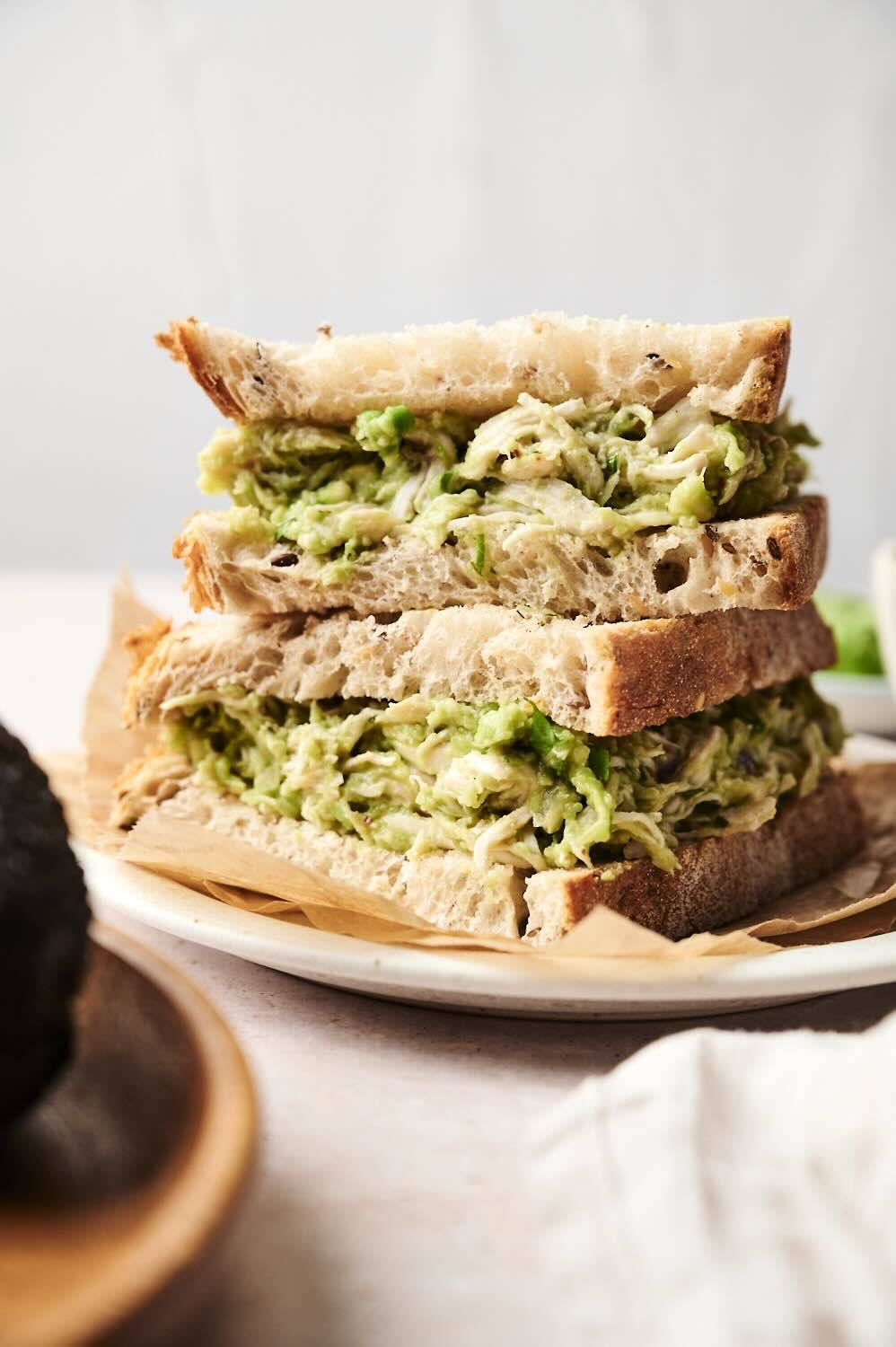Close-up of stacked sandwiches made with creamy avocado chicken salad topped with thick rustic bread. The filling was filled with chopped chicken and mashed potatoes and topped on a white plate with parchment paper.