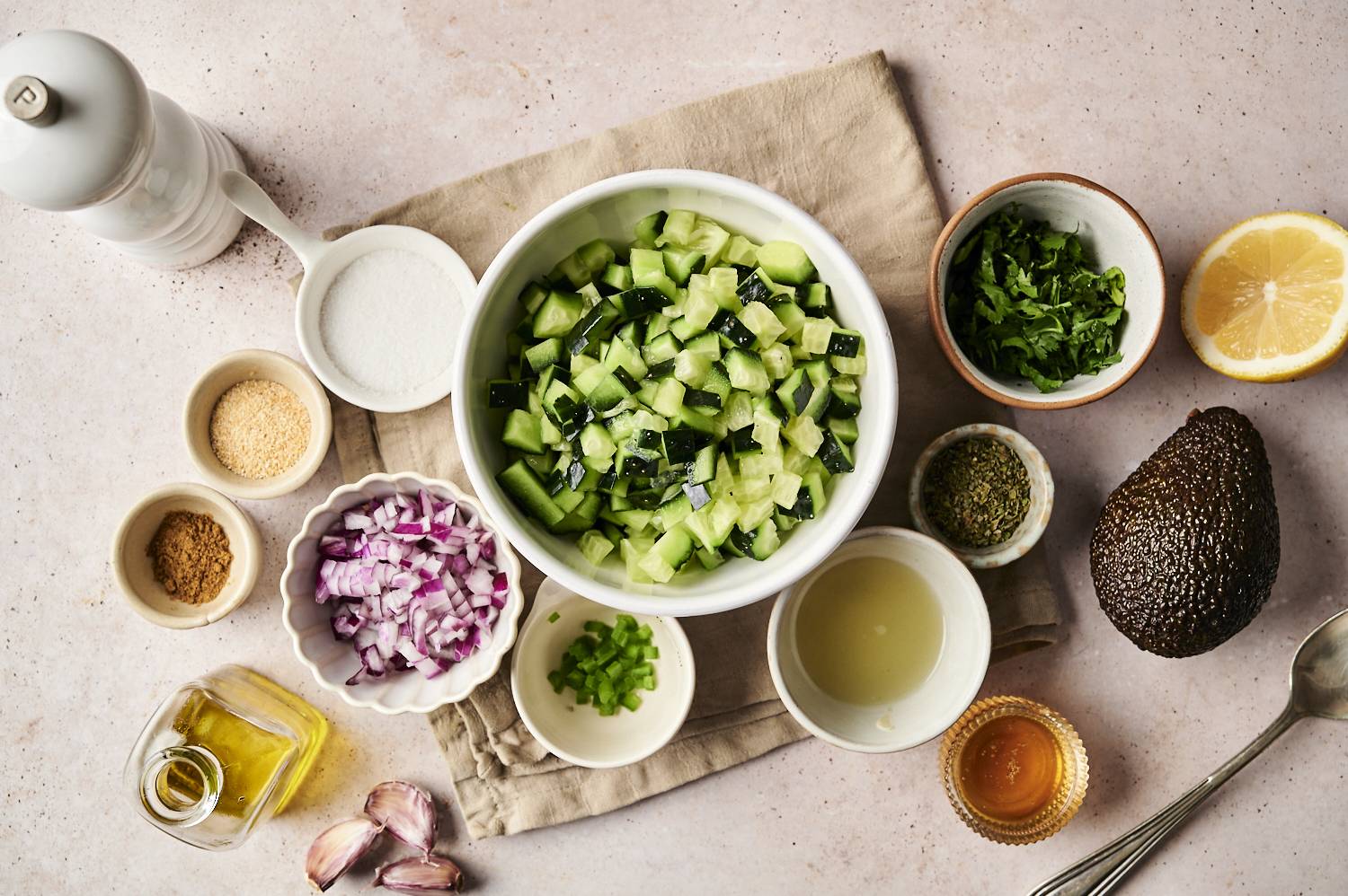 Overhead view of cucumbers diced in a white bowl surrounded by ingredients of avocado cucumber salsa including red onion, jalapeno, coriander, garlic, lemon, lemon, avocado, spices, olive oil, olive oil, olive oil, and salt.
