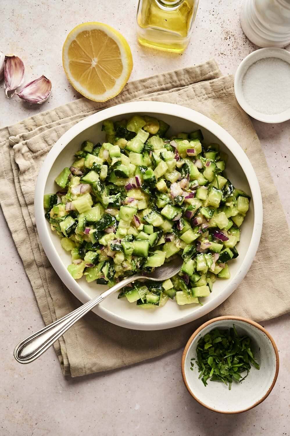 Avocado cucumber salsa top view in white bowl with spoons surrounded by lemon, garlic, olive oil, salt and fresh coriander and on a light-colored surface.