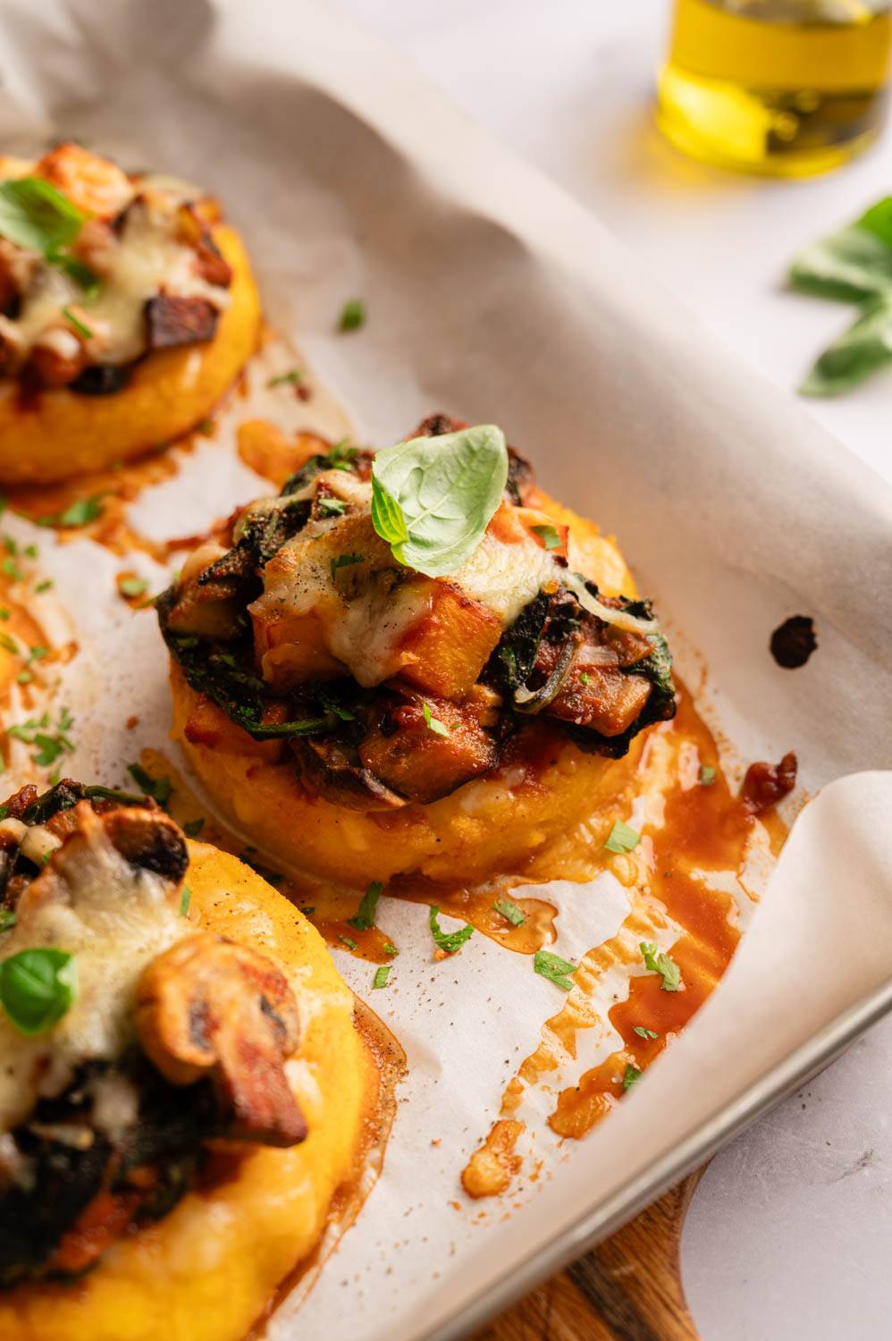 Golden polenta rounds topped with sautéed vegetables, melted cheese, and fresh basil on parchment paper.