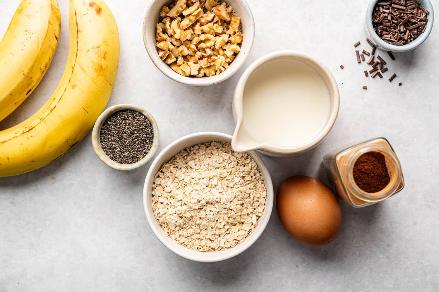 Top view of a kitchen counter filled with ingredients: banana, oats, chopped nuts, chia seeds, chocolate powder, milk, eggs and a jar of cinnamon.