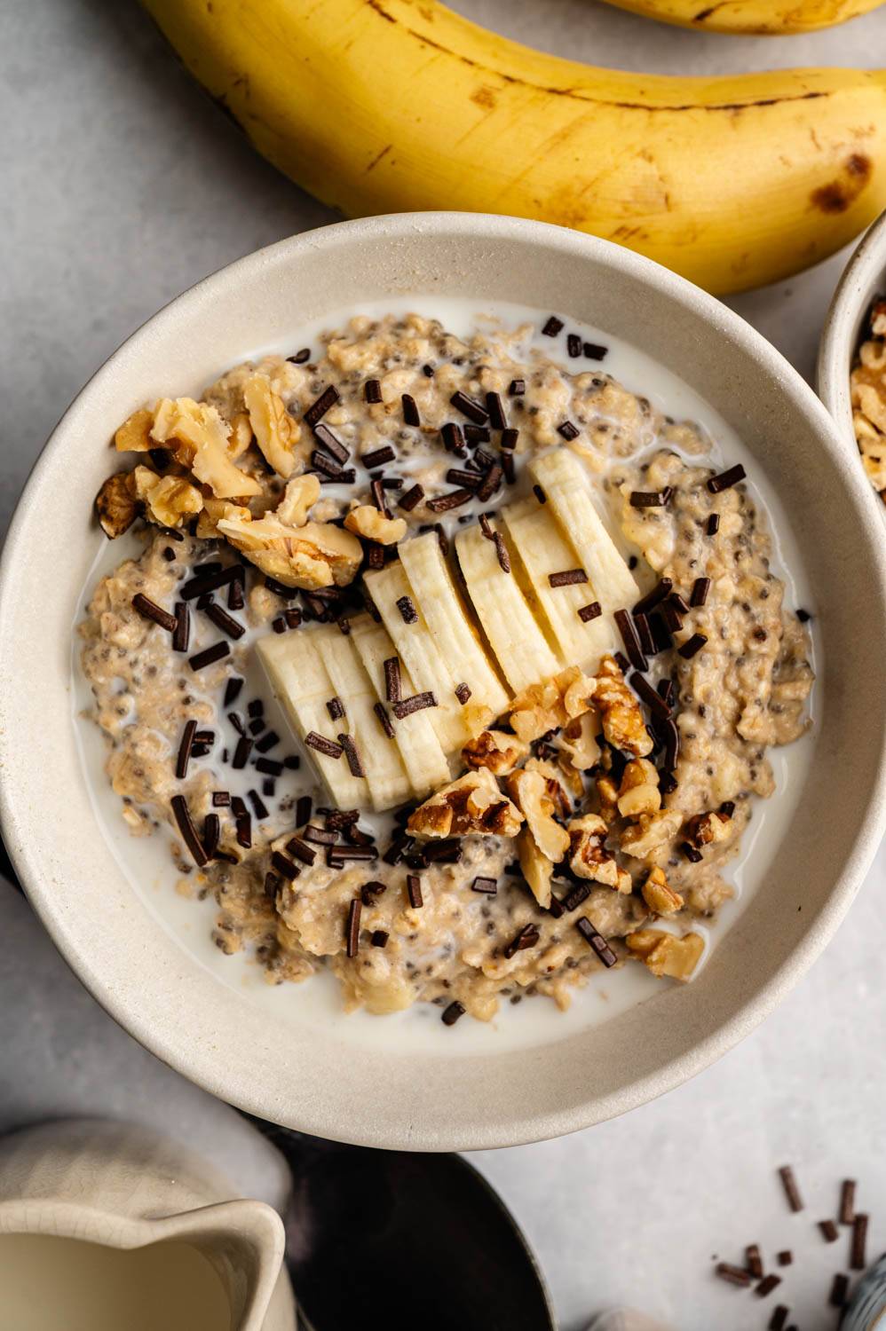 A bowl of oatmeal topped with sliced ​​bananas, walnuts, and chocolate chips is placed next to a whole banana on a light surface to create a cozy, inviting scene.