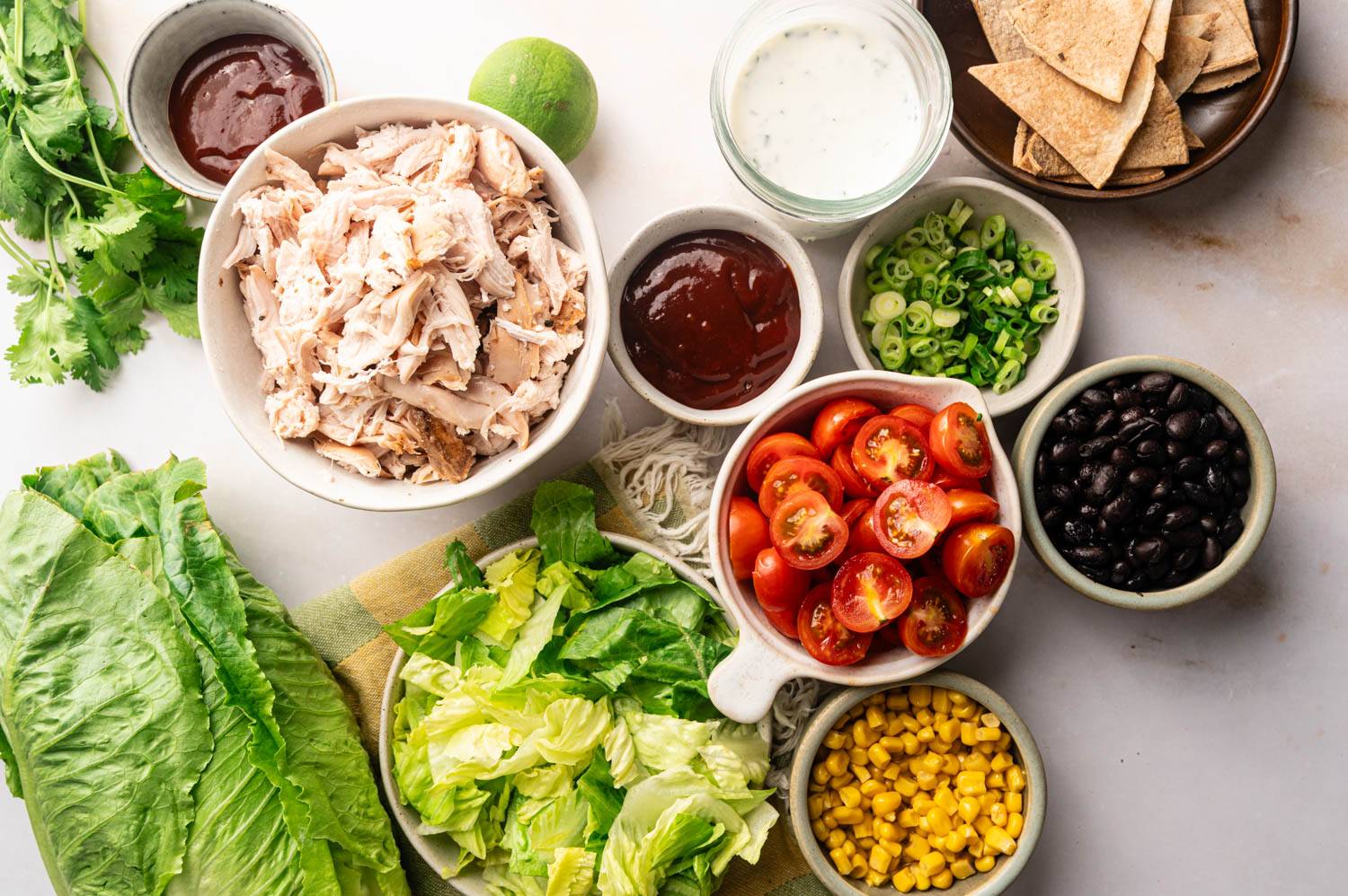 Ingredients for BBQ chicken chopped salad arranged on a countertop, including chopped romaine, shredded chicken, cherry tomatoes, black beans, corn, green onions, cilantro, ranch dressing, barbecue sauce, lime, and tortilla chips.