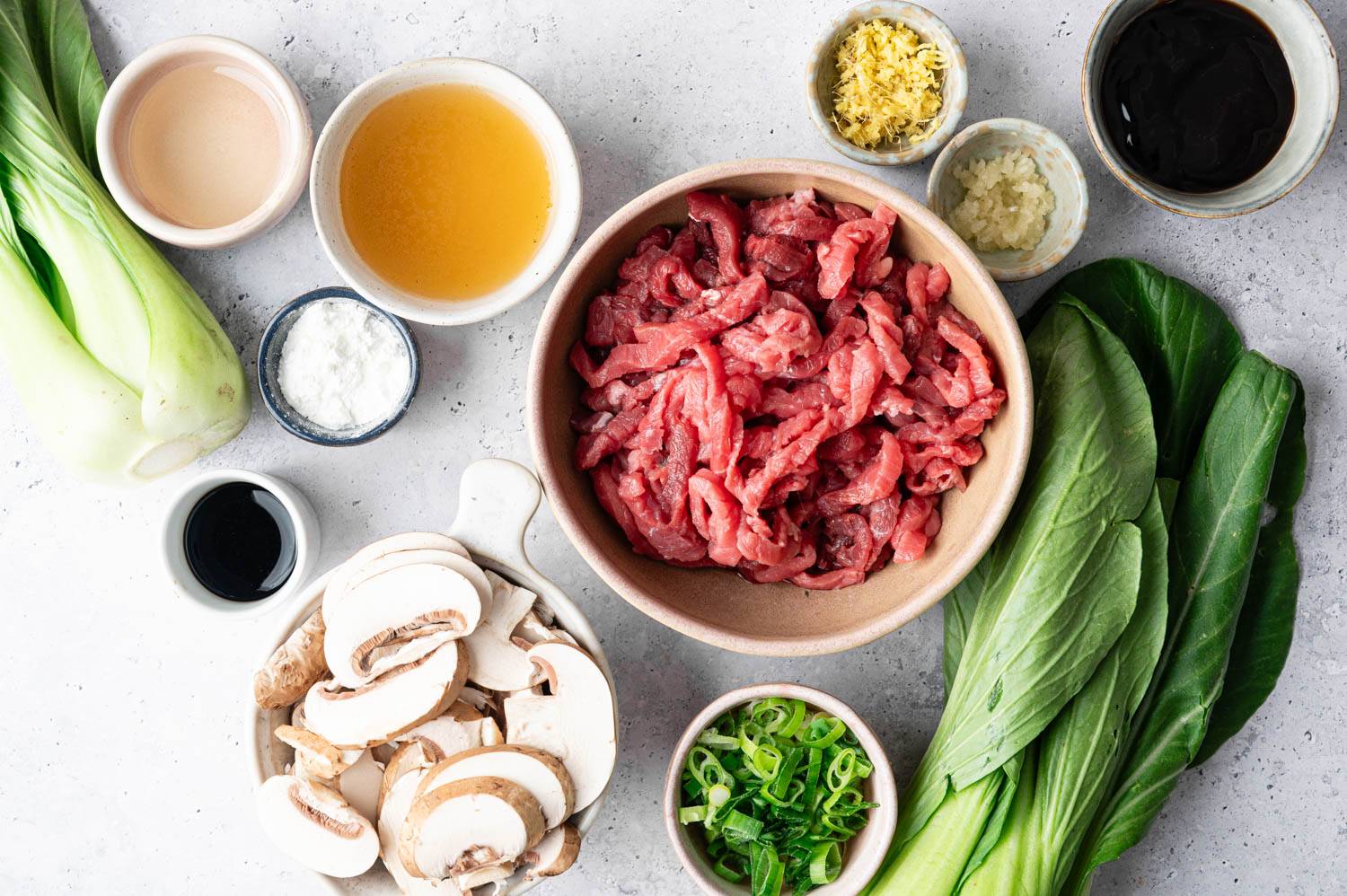 A beautifully arranged assortment of ingredients on a light surface includes bok choy, sliced raw beef, mushrooms, green onions, minced ginger, garlic, and various sauces in bowls, conveying a fresh and inviting culinary preparation scene.