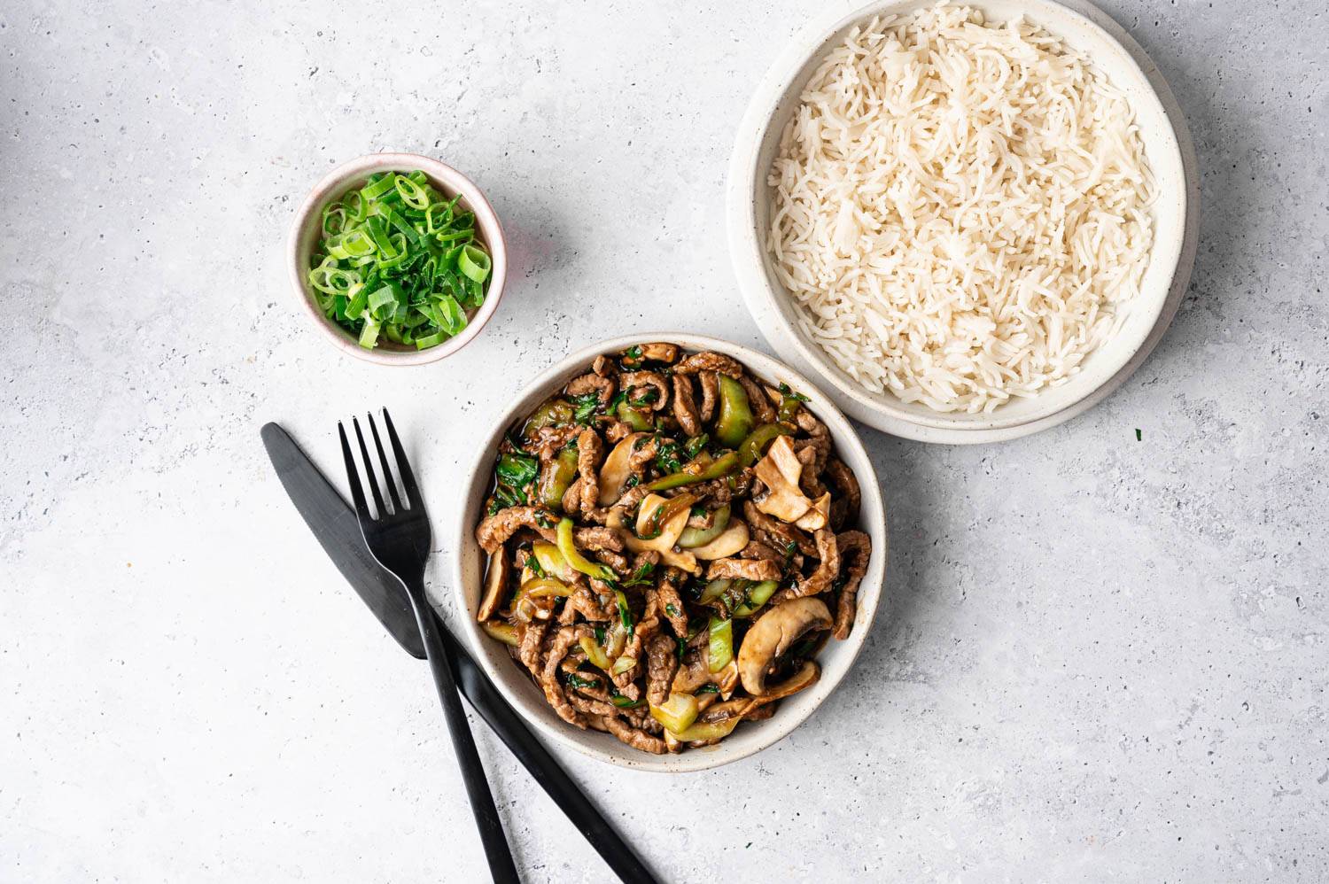 A bowl of stir-fried beef with mushrooms and green peppers is accompanied by a bowl of white rice. A small dish of chopped green onions is beside them, with a black fork and knife on the side.