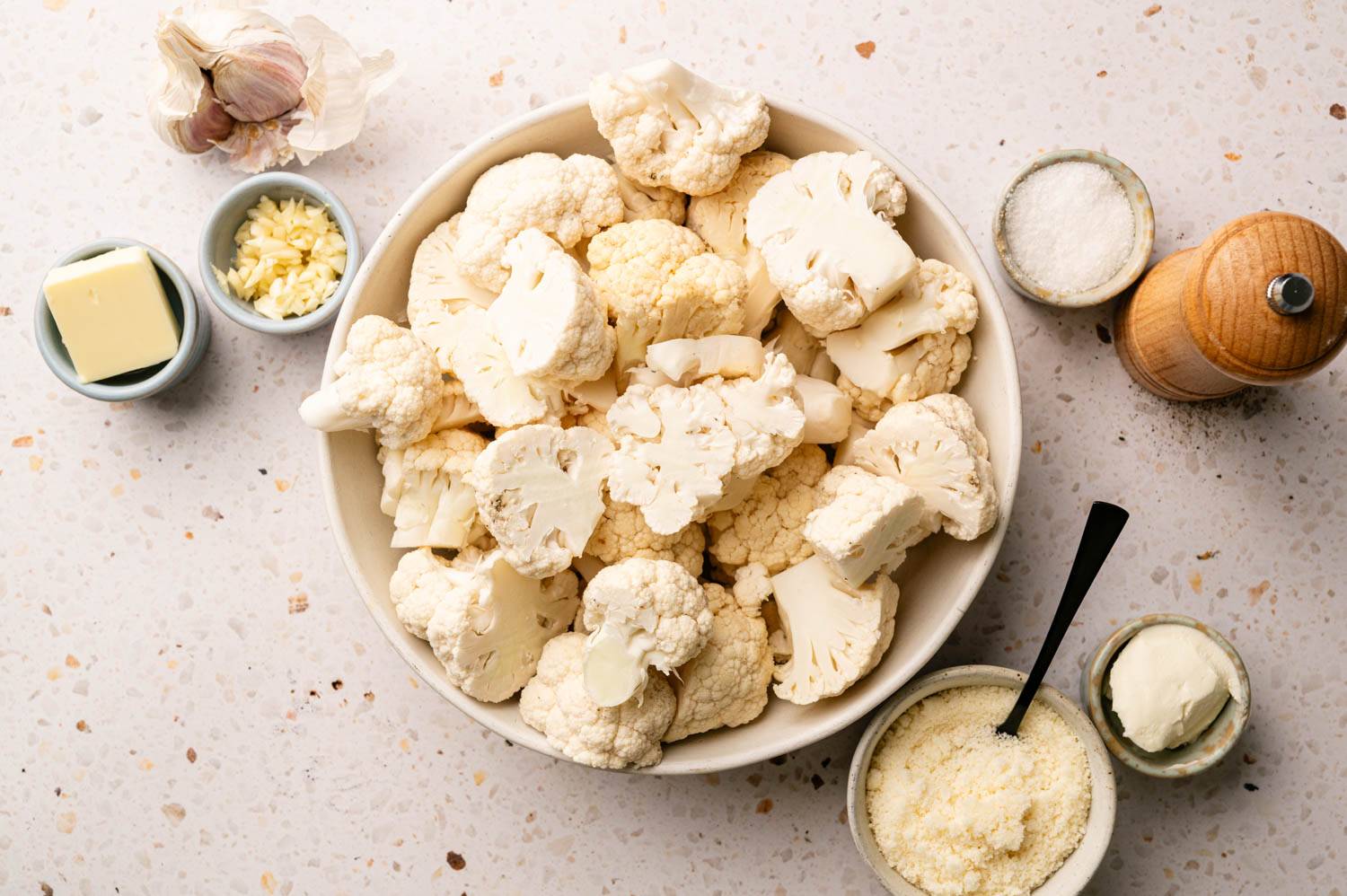 Bowl of fresh cauliflower florets on a speckled countertop, surrounded by garlic cloves, butter, salt, and cheese.