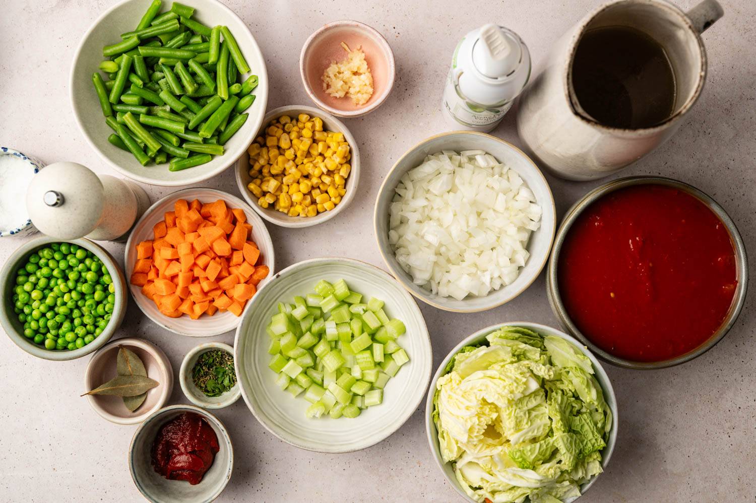 Assorted bowls of vegetables for soup, including chopped carrots, onions, celery, peas, and cabbage, surround a pitcher and canned tomatoes, evoking freshness.