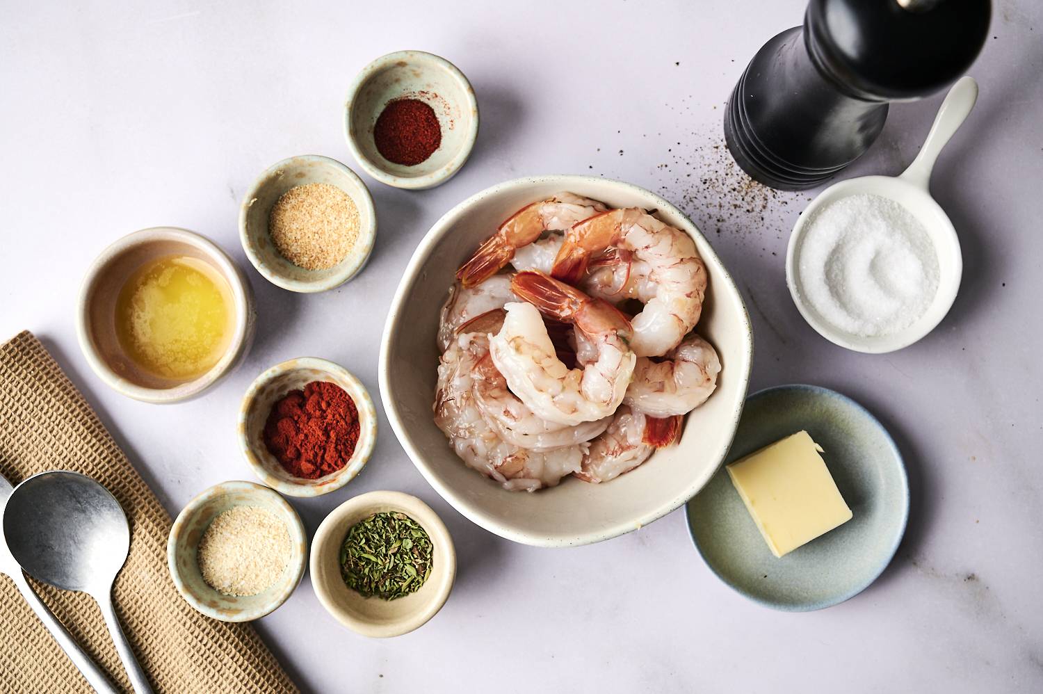A bowl of raw shrimp is surrounded by a variety of spices, oil, butter and pepper grinders on a light countertop.