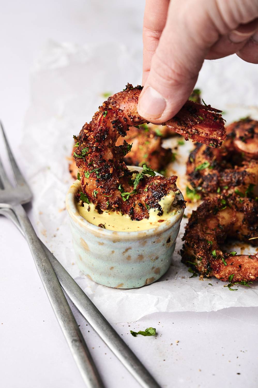 One hand dipped crispy seasoned shrimp in a small bowl of creamy sauce with more shrimp and parsley on the background.