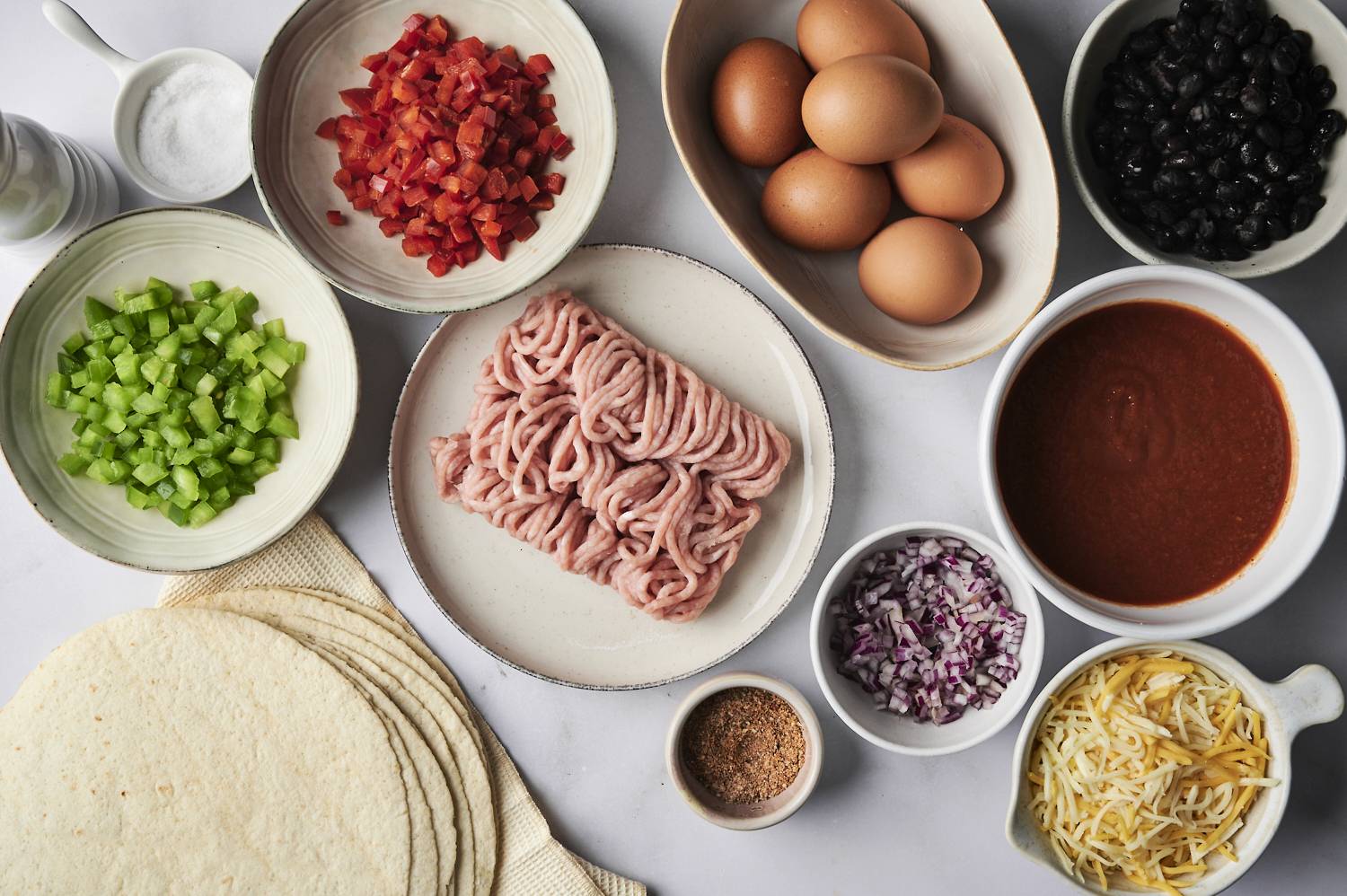 The ingredients for breakfast enchiladas are displayed: diced red bell peppers, diced green bell peppers, ground turkey, eggs, black beans, enchilada sauce, red onions, taco seasoning, shredded cheese, and tortillas.