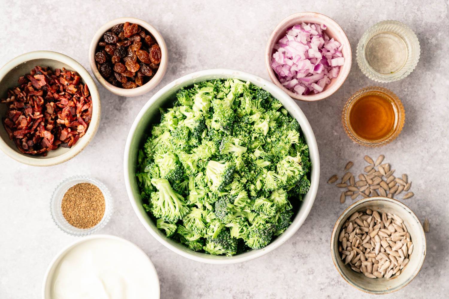 A bowl of fresh broccoli florets is surrounded by ingredients: sunflower seeds, raisins, chopped onion, crispy bacon, yogurt, vinegar, and mustard seeds.
