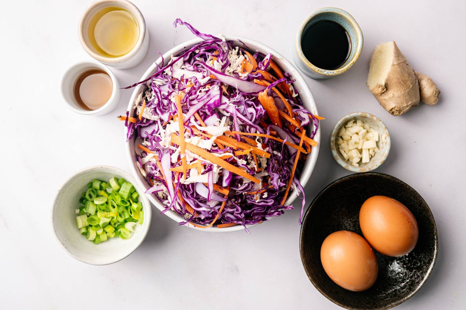 A bowl of chopped purple cabbage and carrots surrounded by green onions, eggs, chopped garlic, soy sauce, ginger, vinegar and oil on a white surface.