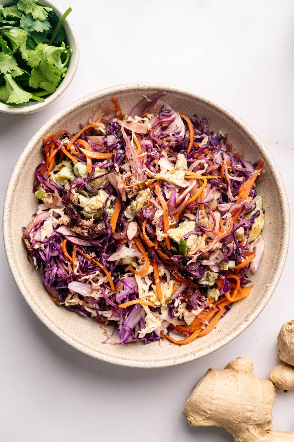 Vibrant salad with chopped red cabbage, carrots and green leafy vegetables in a beige bowl. A bowl of cilantro and fresh ginger root is nearby.