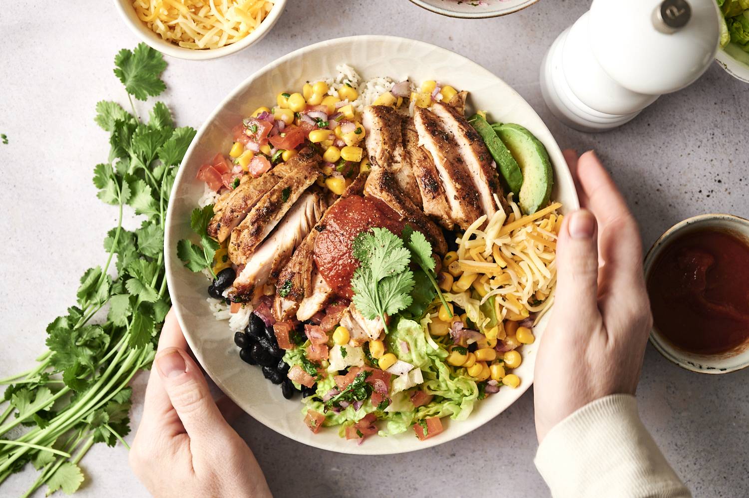 One hand holds a bowl of colorful chicken burritos with rice, black beans, corn, avocado, cheese and salsa, garnished with cilantro.