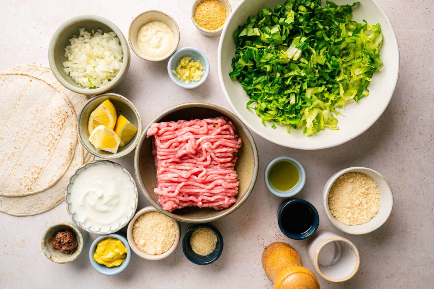 Overhead view of chicken Caesar smash taco ingredients in small bowls, including raw ground chicken, chopped romaine, diced onion, lemon wedges, Greek yogurt, Parmesan, anchovy paste, Dijon, and a stack of flour tortillas.
