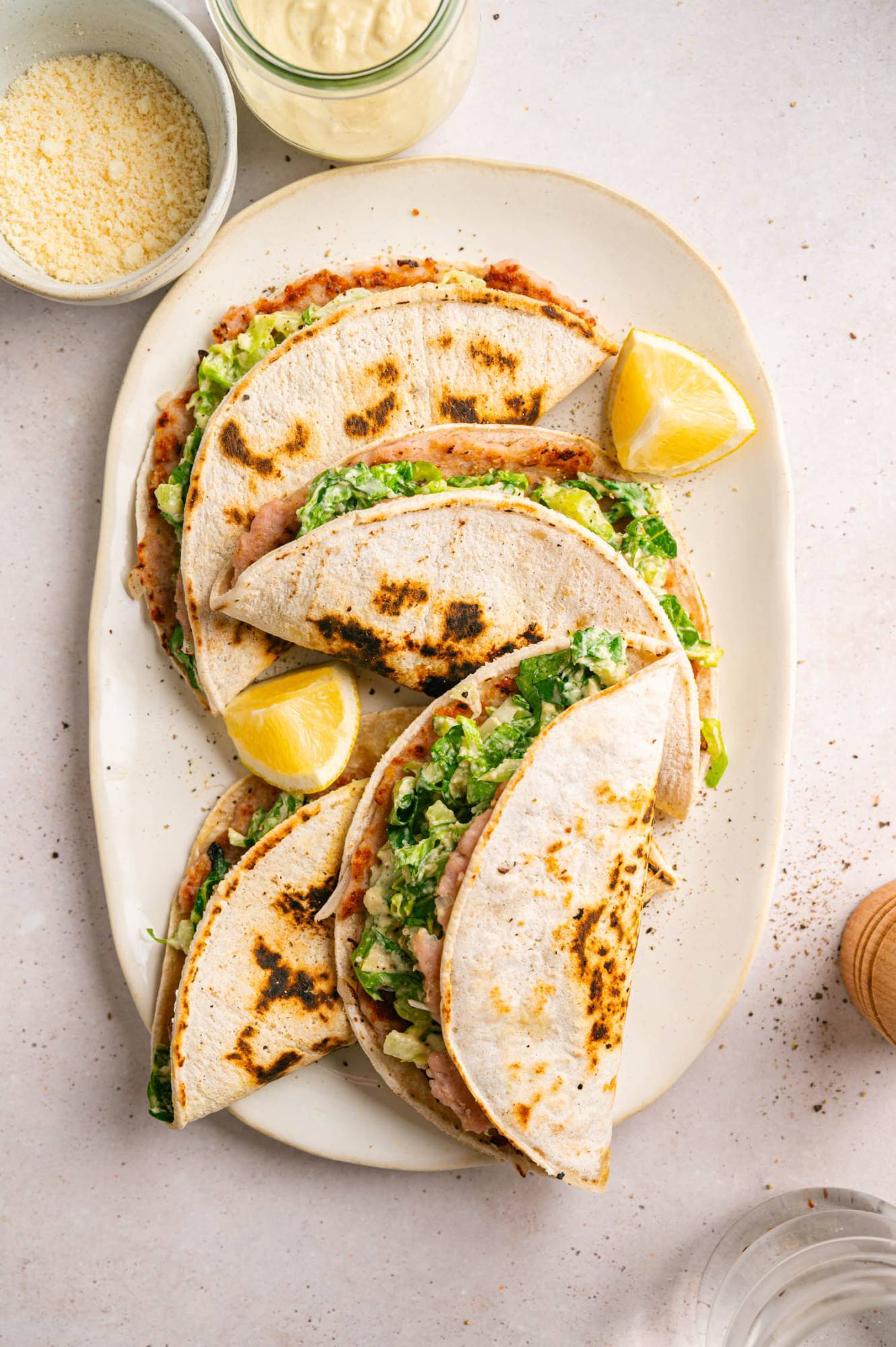 Overhead view of chicken Caesar smash tacos folded on an oval platter with lemon wedges and small bowls of Parmesan and Caesar dressing.
