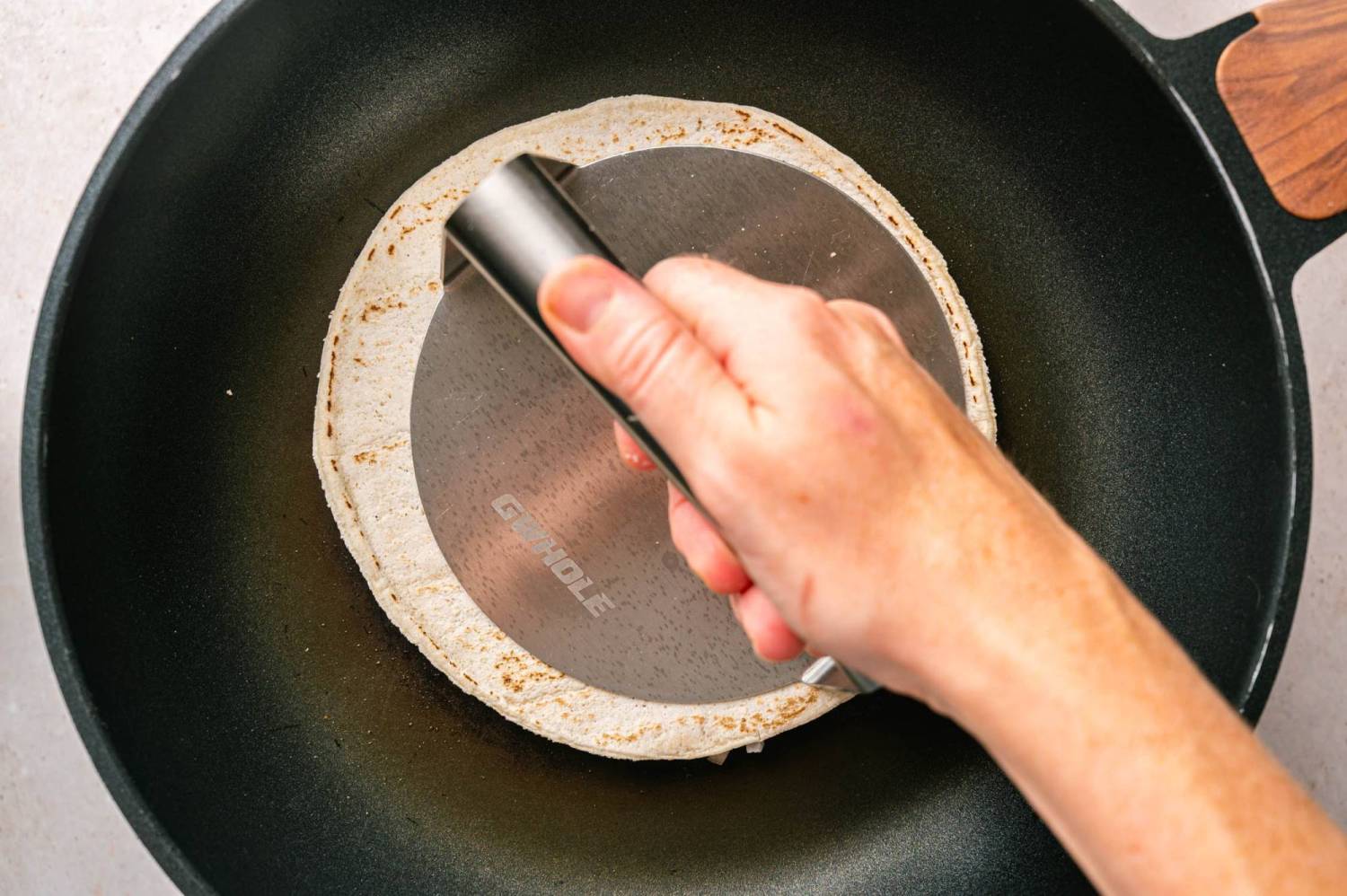 Hand pressing a flour tortilla down onto a ball of ground chicken in a hot skillet with a stainless steel burger smasher.