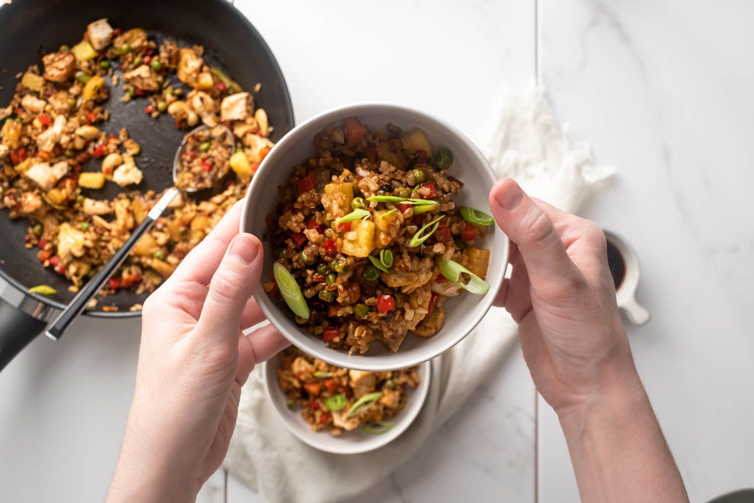 Chicken fried rice with pineapple, cashews, green onions and vegetables in a bowl held by two hands.