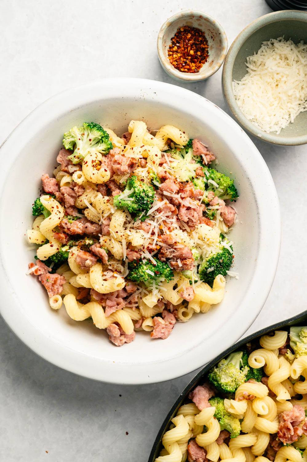 A bowl of curly pasta topped with sausage, broccoli, and grated cheese is served with a few bowls of red pepper flakes and Parmesan cheese for a delicious, savory appetizer.