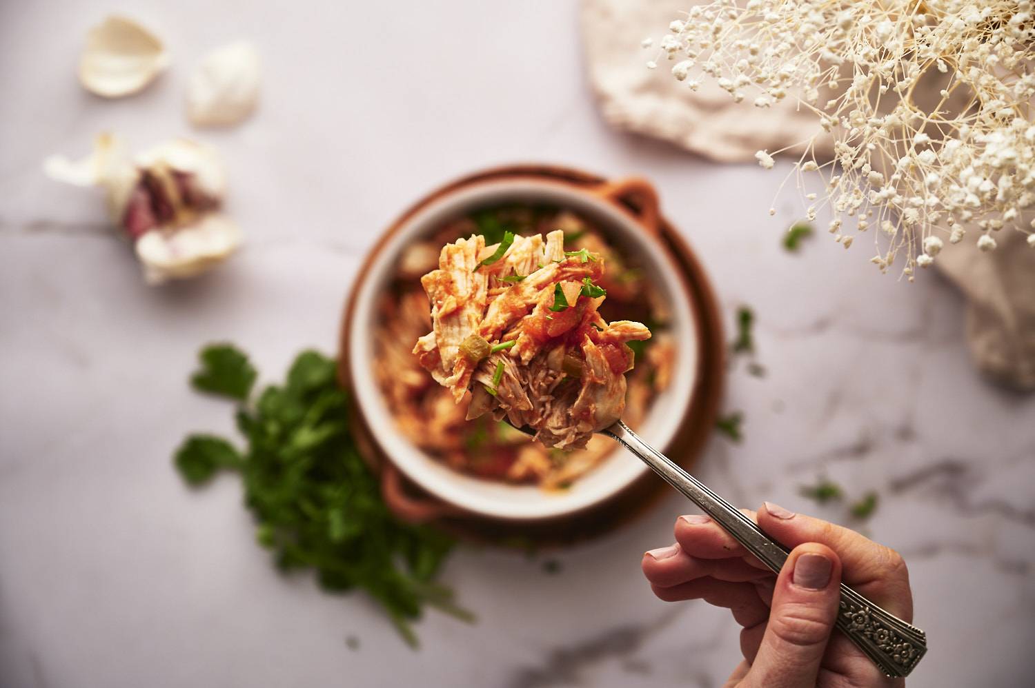 In one hand, chopped chicken with a fork, mixed with tomatoes and herbs, on a bowl on a marble surface, with herbs nearby.