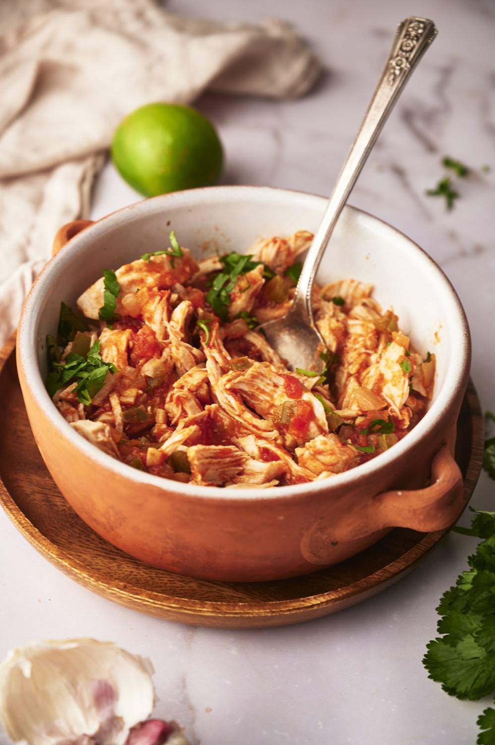A bowl of thinly sliced ​​chicken mixed with tomatoes and herbs, topped with cilantro and next to lime and garlic on the marble surface.