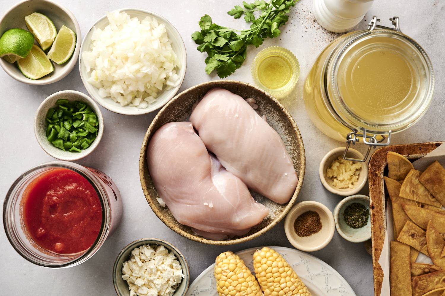 Ingredients for chicken tortilla soup laid out, including raw chicken breasts, corn, diced onions, lime wedges, cilantro, tomato sauce, chicken broth, and seasonings.