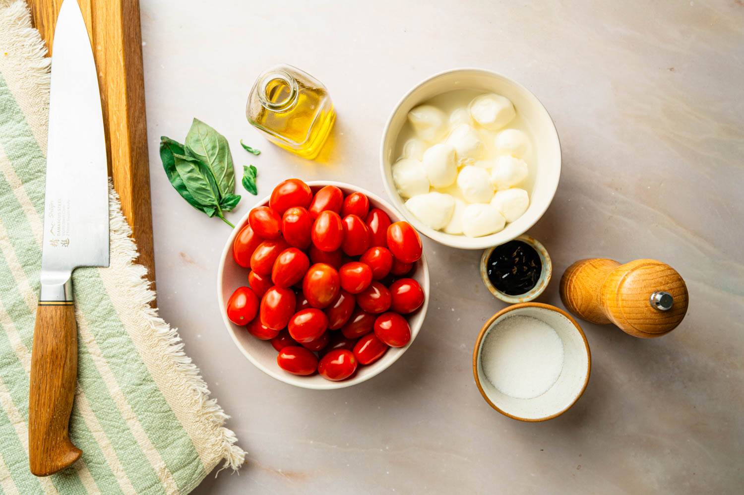 Ingredients for chopped Caprese salad including cherry tomatoes, fresh mozzarella, basil, olive oil, balsamic glaze, salt, and pepper on a kitchen counter.