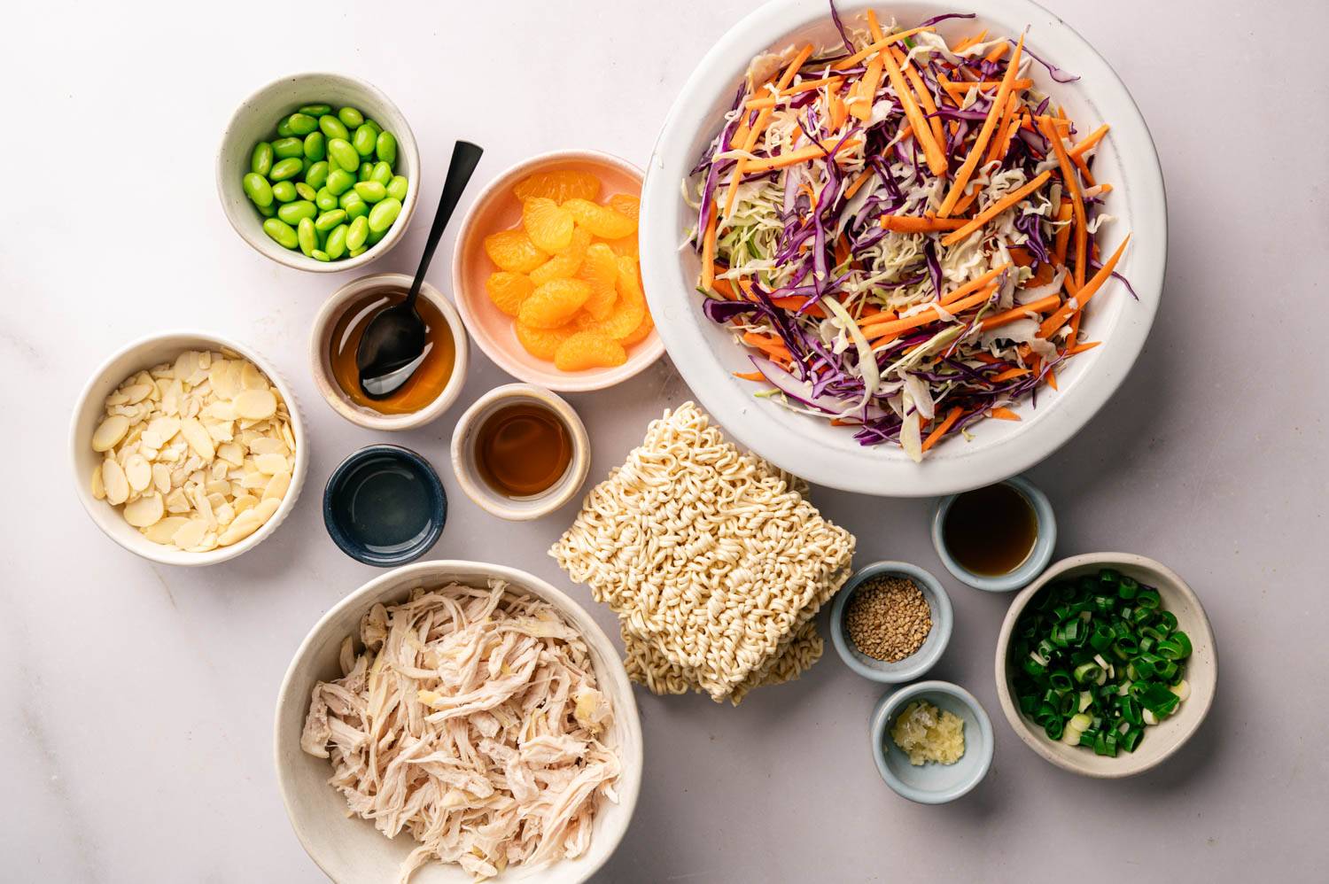 Ingredients for a vibrant Asian salad: chopped cabbage and carrots, edamame, citrus slices, almonds, chopped chicken, ramen noodles, soy sauce, sesame seeds, scallions and minced garlic.