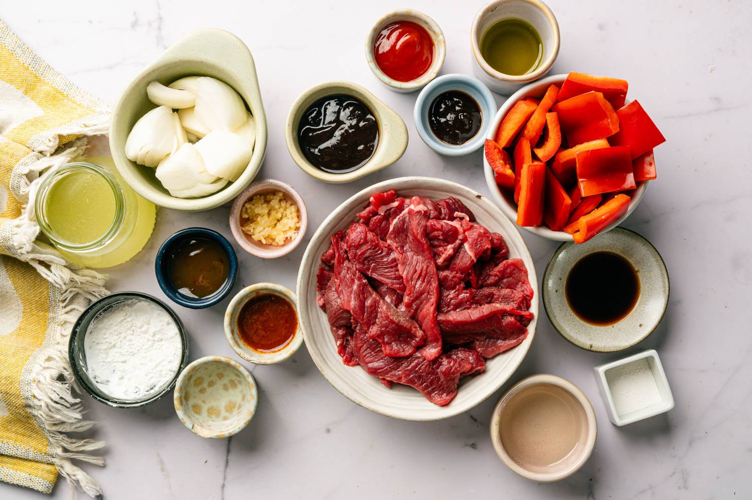 A white marble surface displays ingredients in small bowls: raw beef slices, red bell peppers, chopped onions, sauces, garlic, and seasonings, arranged neatly.